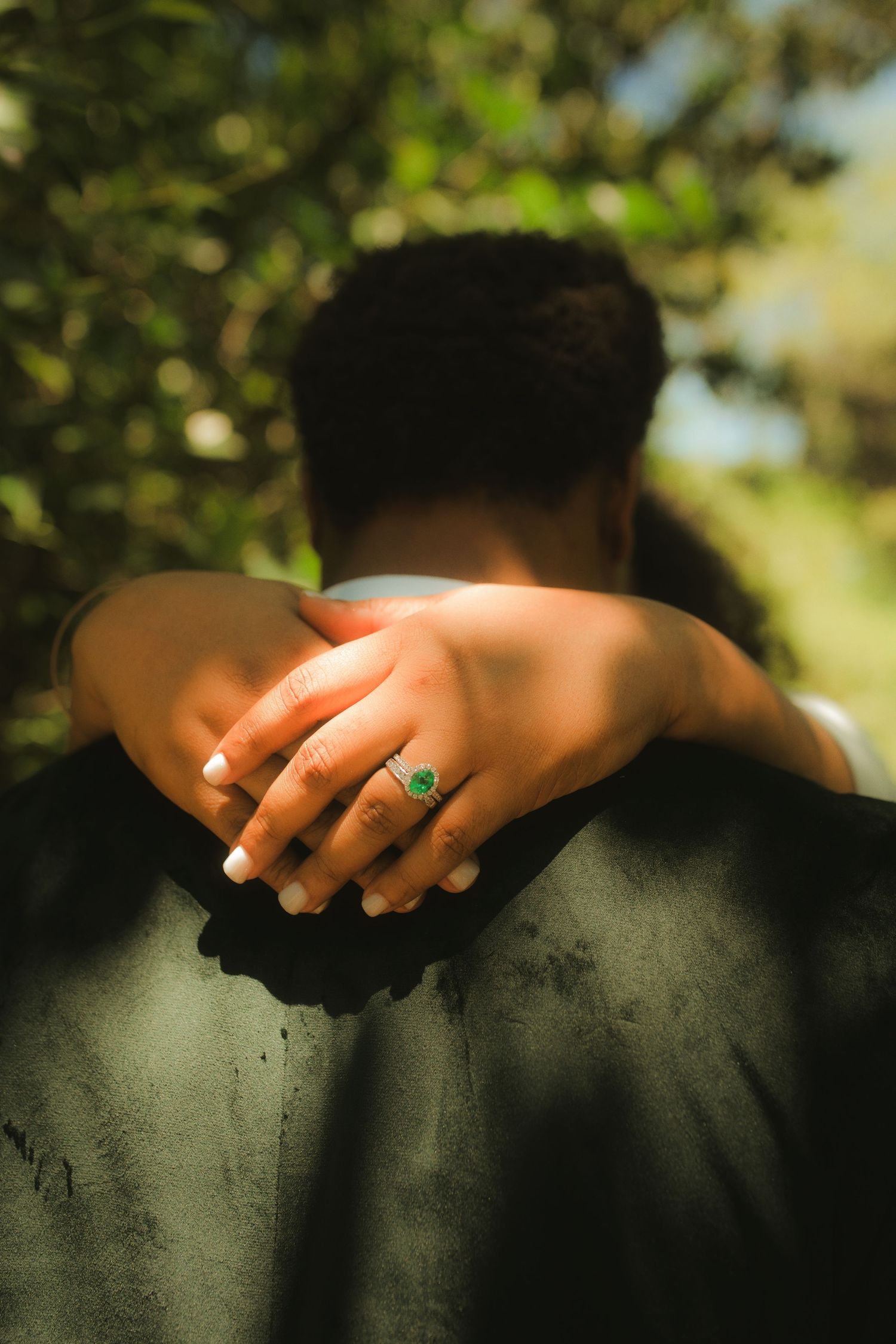 Close-up of bride's hand with green emerald engagement ring resting on groom's dark suit.