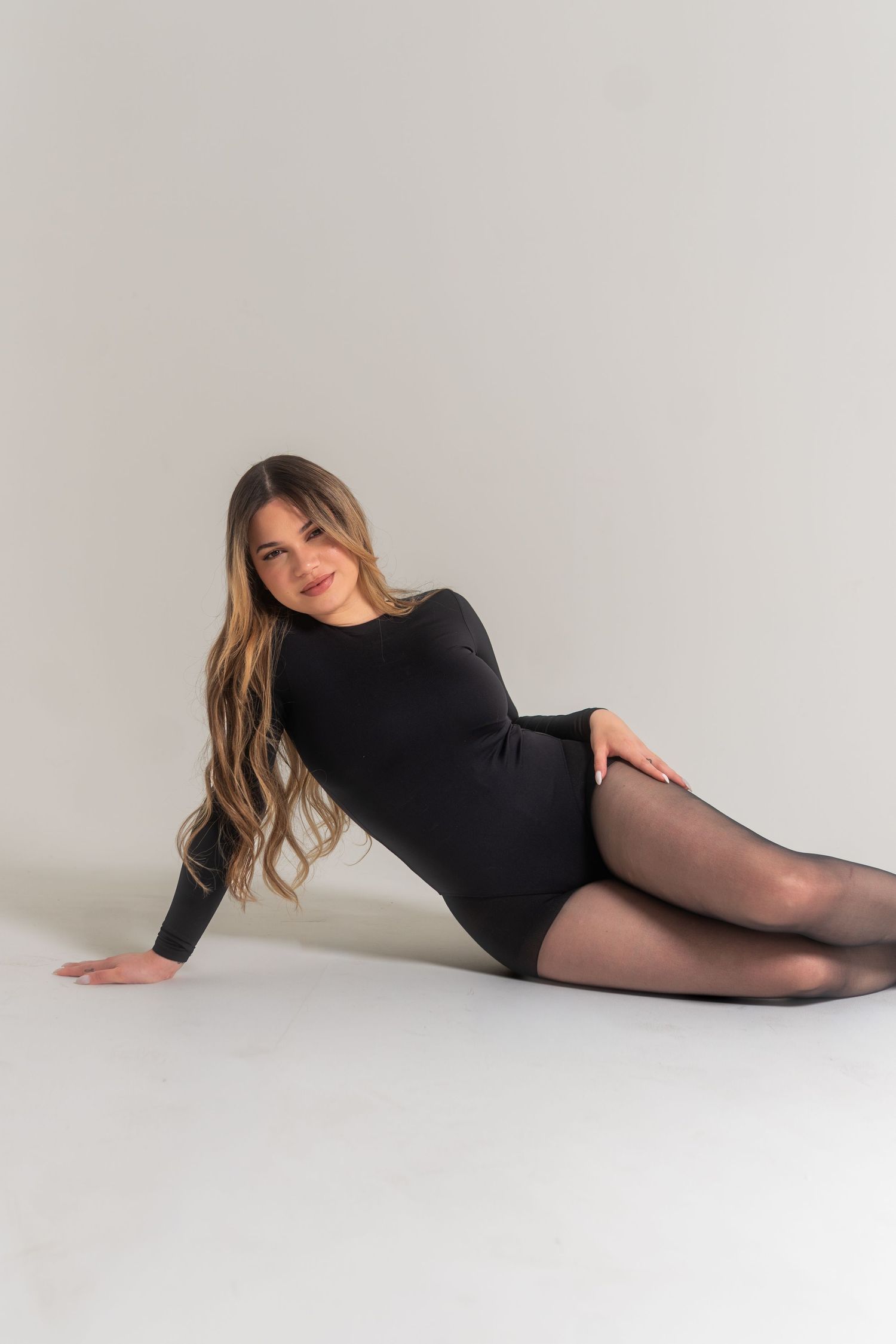 Woman in black long-sleeve bodysuit and sheer tights poses gracefully on a white studio floor.