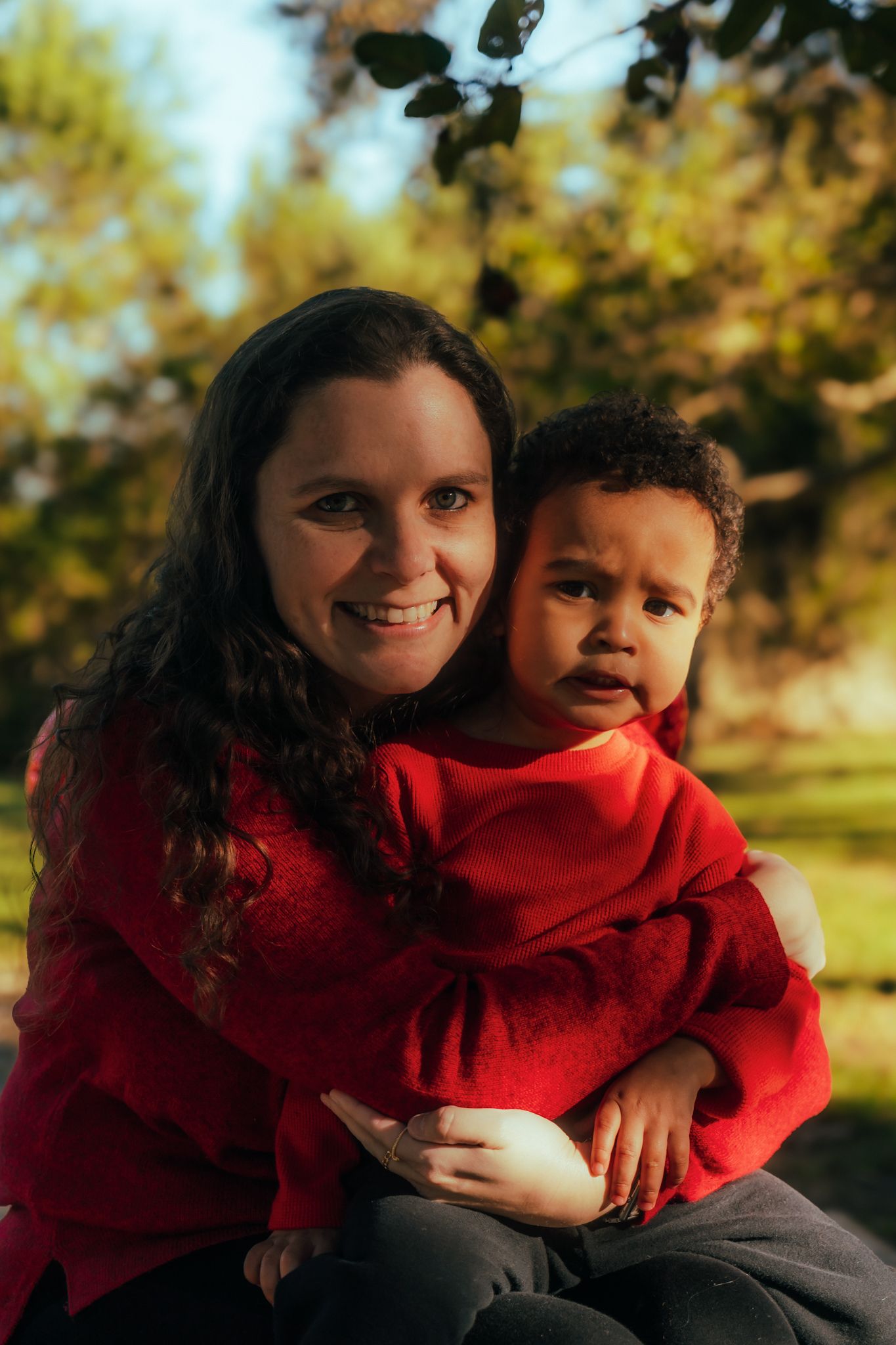 A smiling woman in red holds a young child outdoors, surrounded by lush green trees in warm sunlight.