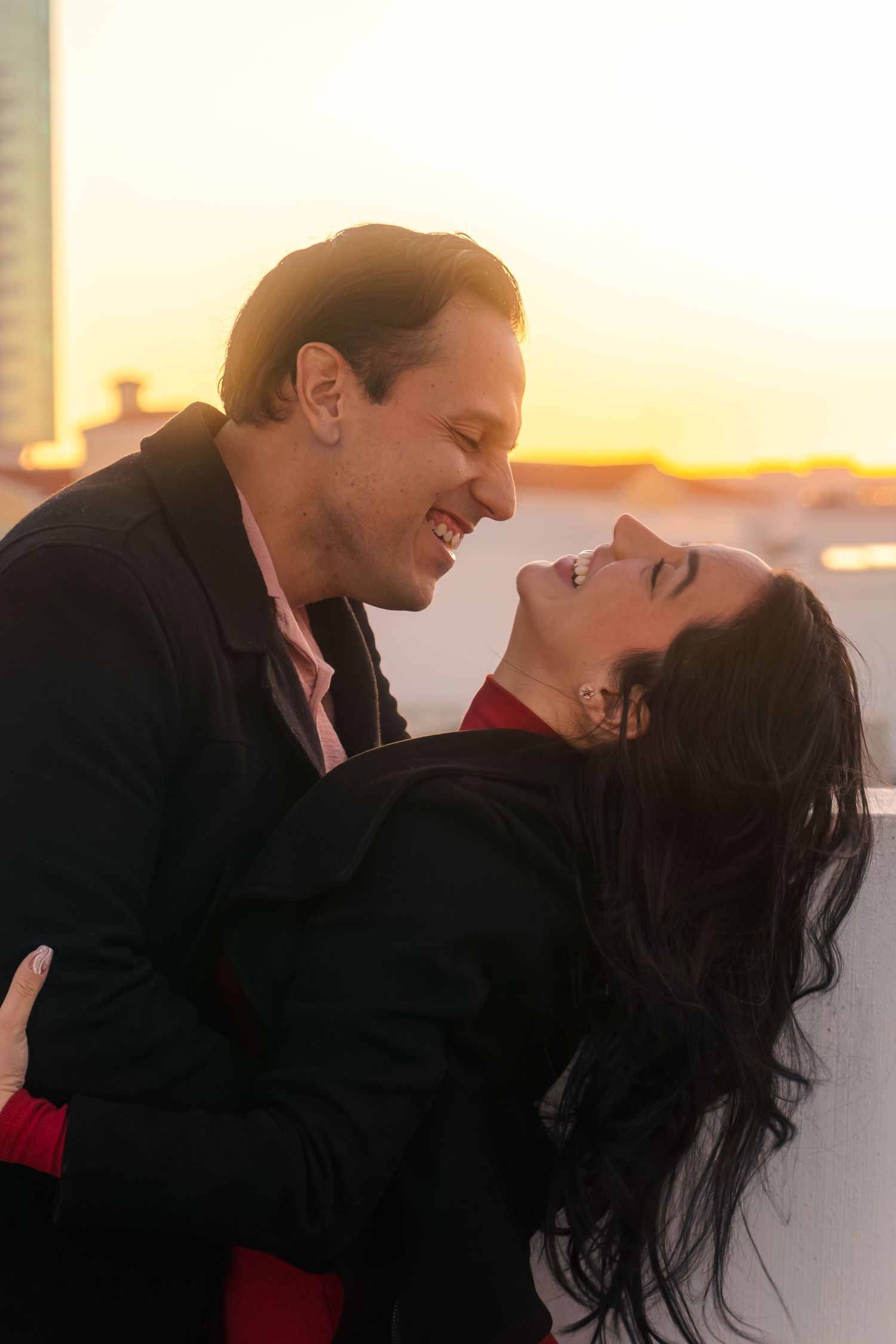Happy couple laughing and embracing on a rooftop at golden hour sunset with city buildings behind.