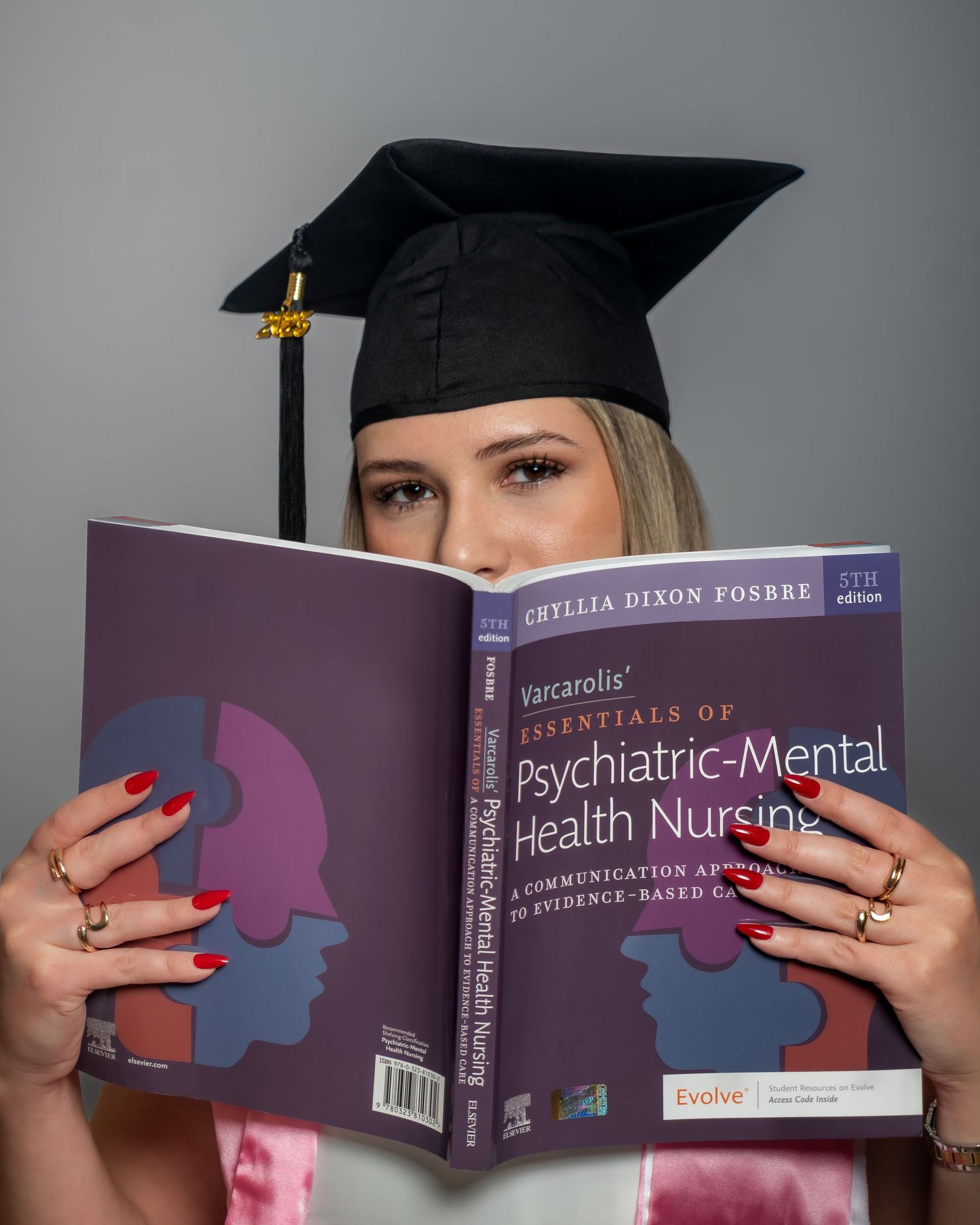 Graduate in cap holds Varcarolis Psychiatric-Mental Health Nursing textbook, wearing red nails and rings.