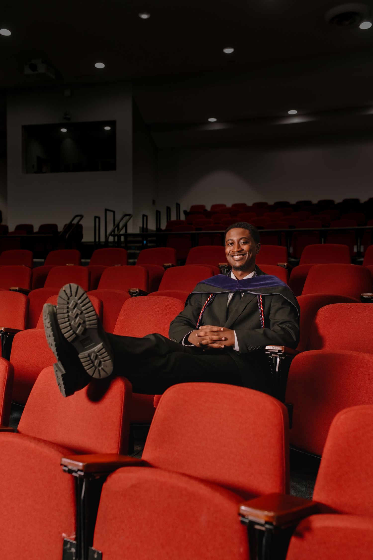 Smiling graduate in cap and gown relaxing in empty red theater seats with feet up.