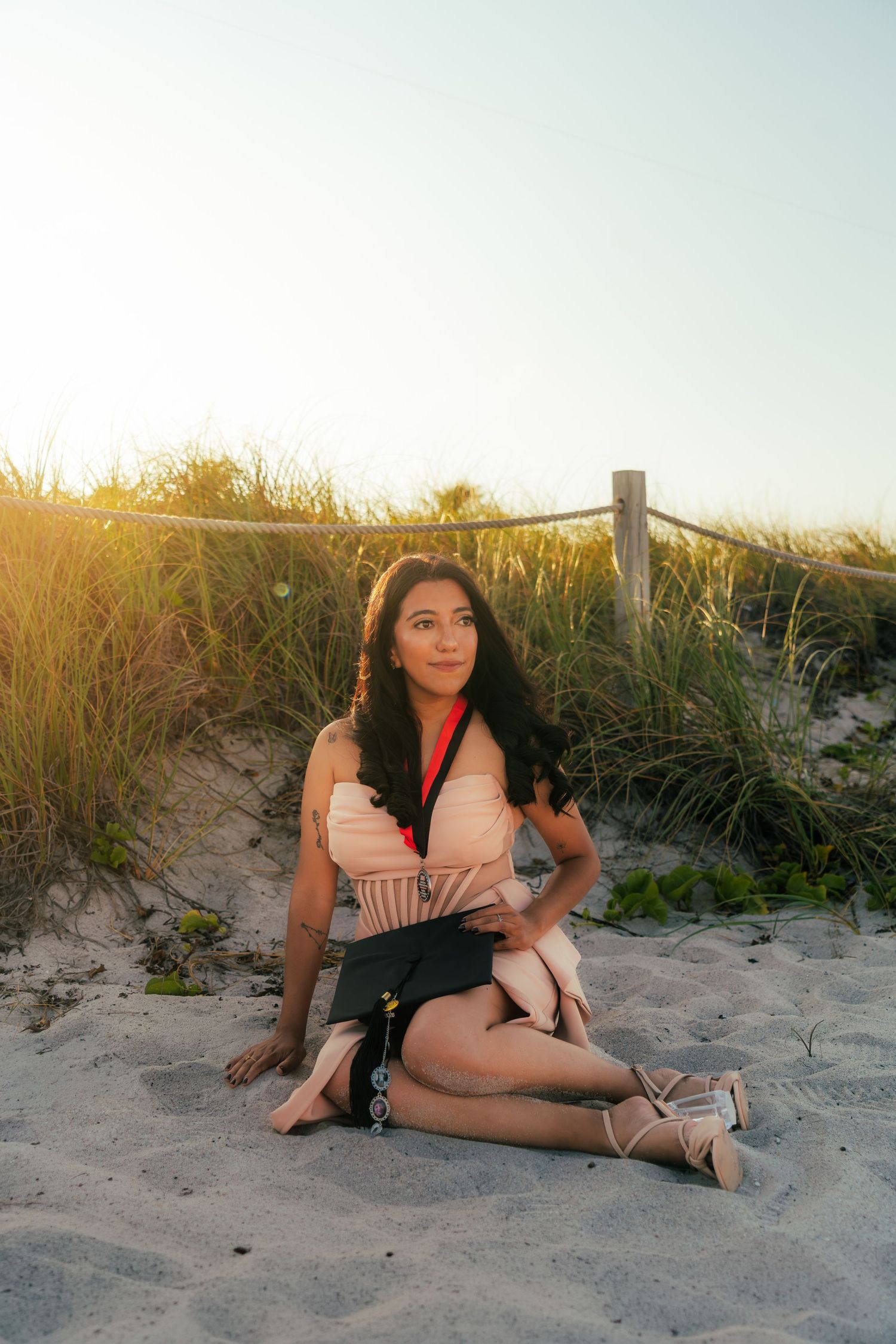 Young woman sitting on sandy beach dunes at golden hour holding a clutch purse.