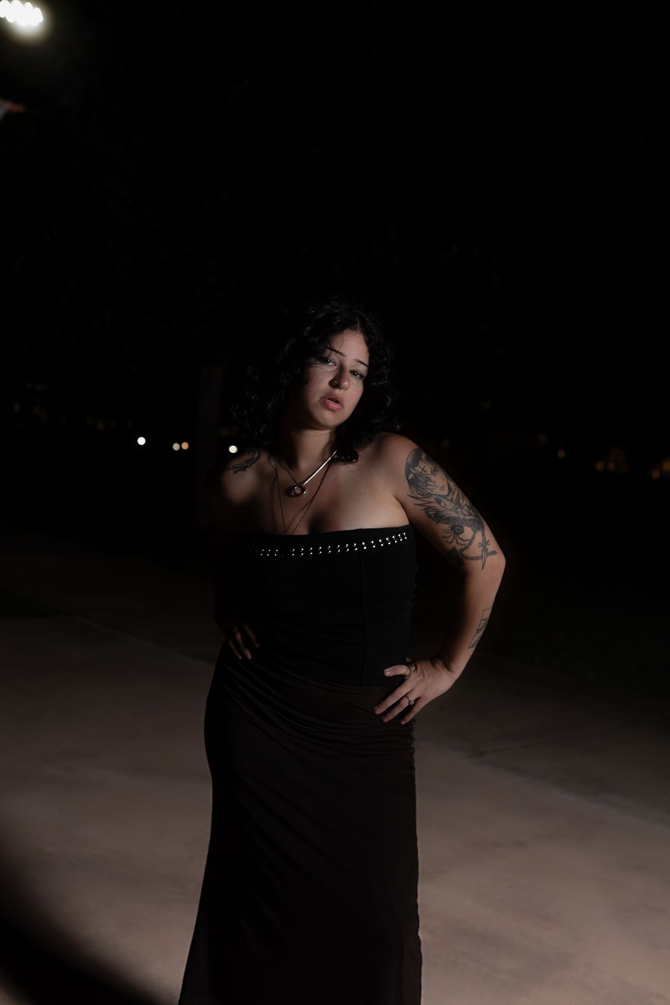 Woman in black strapless gown with tattoo poses confidently outdoors at night.