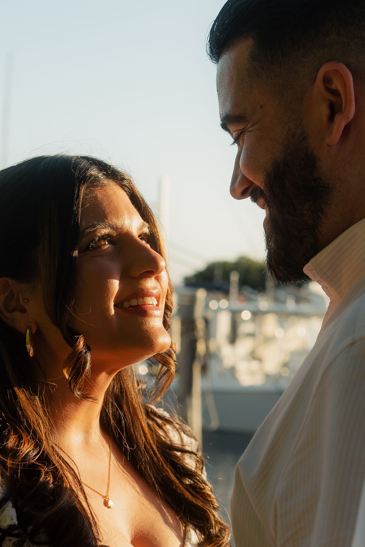 Smiling woman with long dark hair gazes at bearded man during golden hour at marina.
