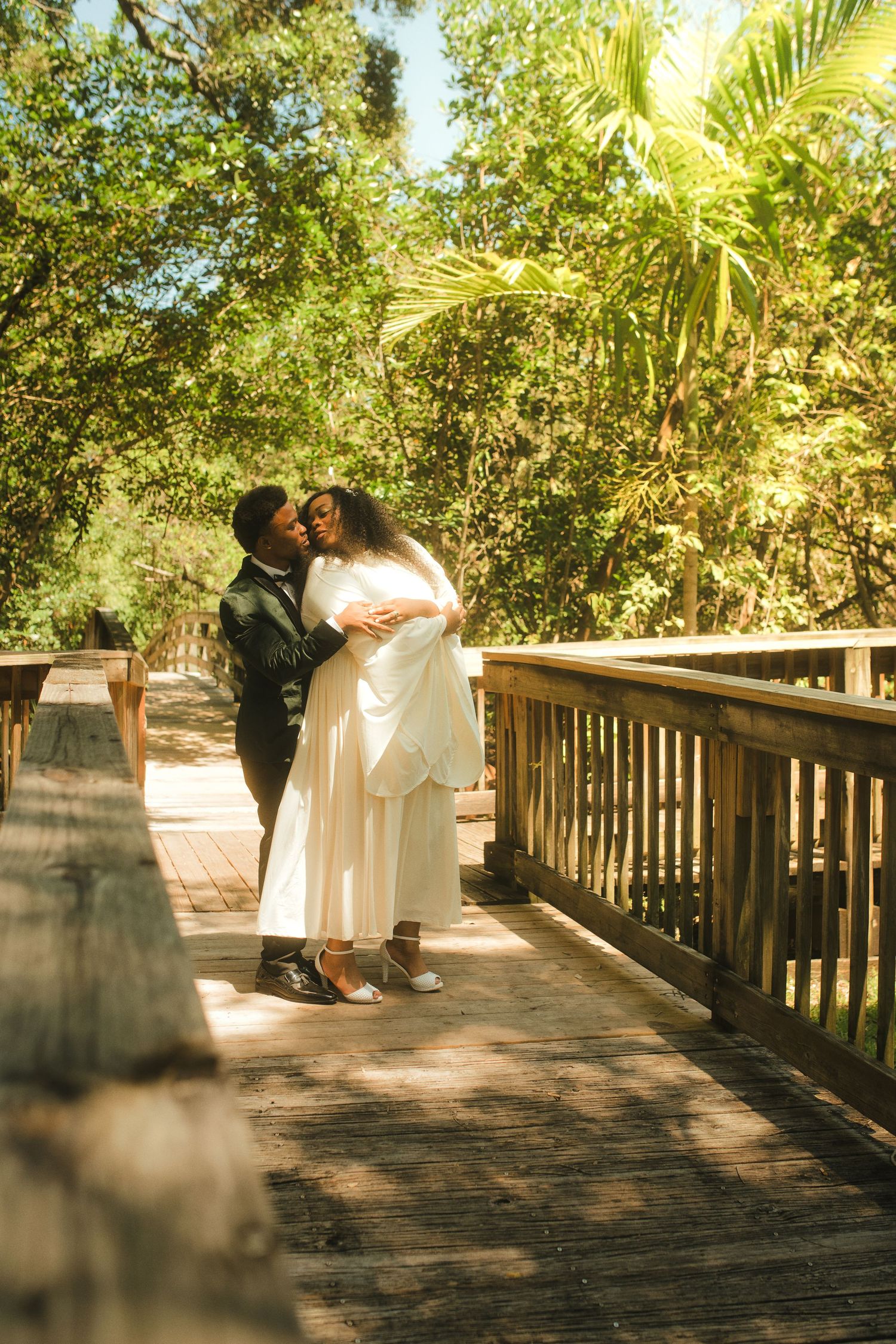 Couple embracing on a sunny wooden boardwalk surrounded by lush green trees, woman in white dress and man in dark jacket.