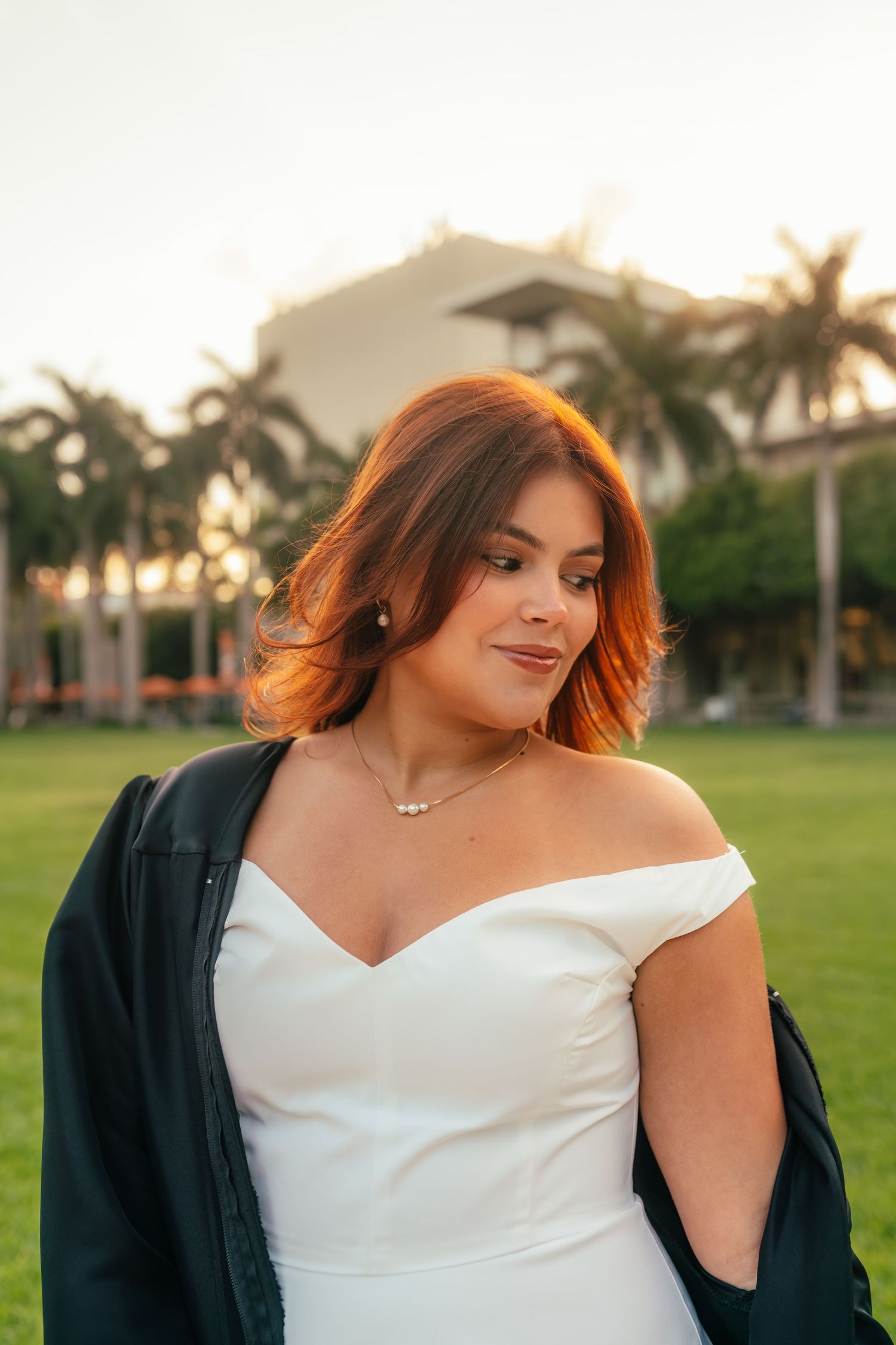 Woman in white off-shoulder dress and black jacket poses outdoors at golden hour near palm trees.