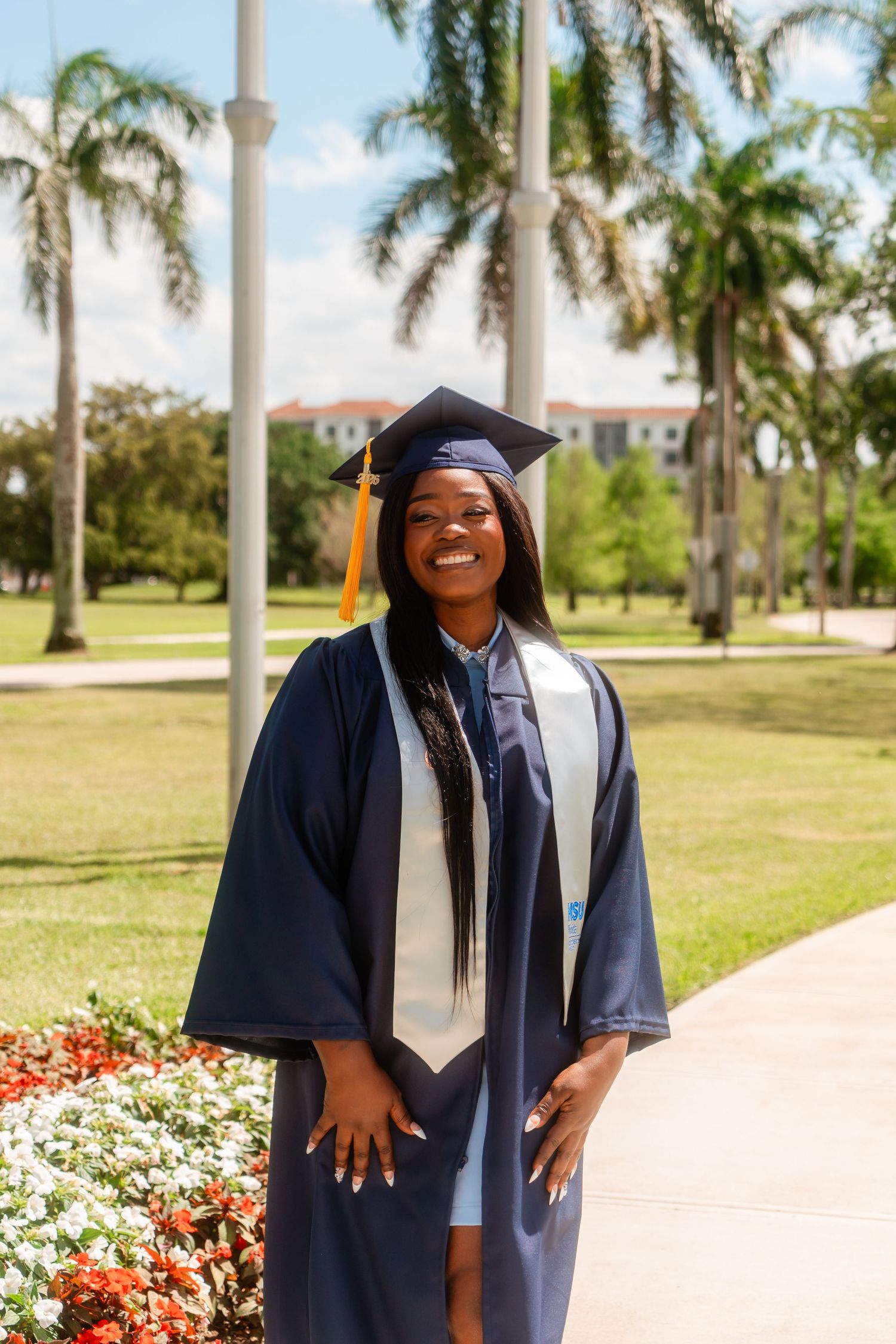 Smiling graduate in navy cap and gown stands by colorful flowers and palm trees.