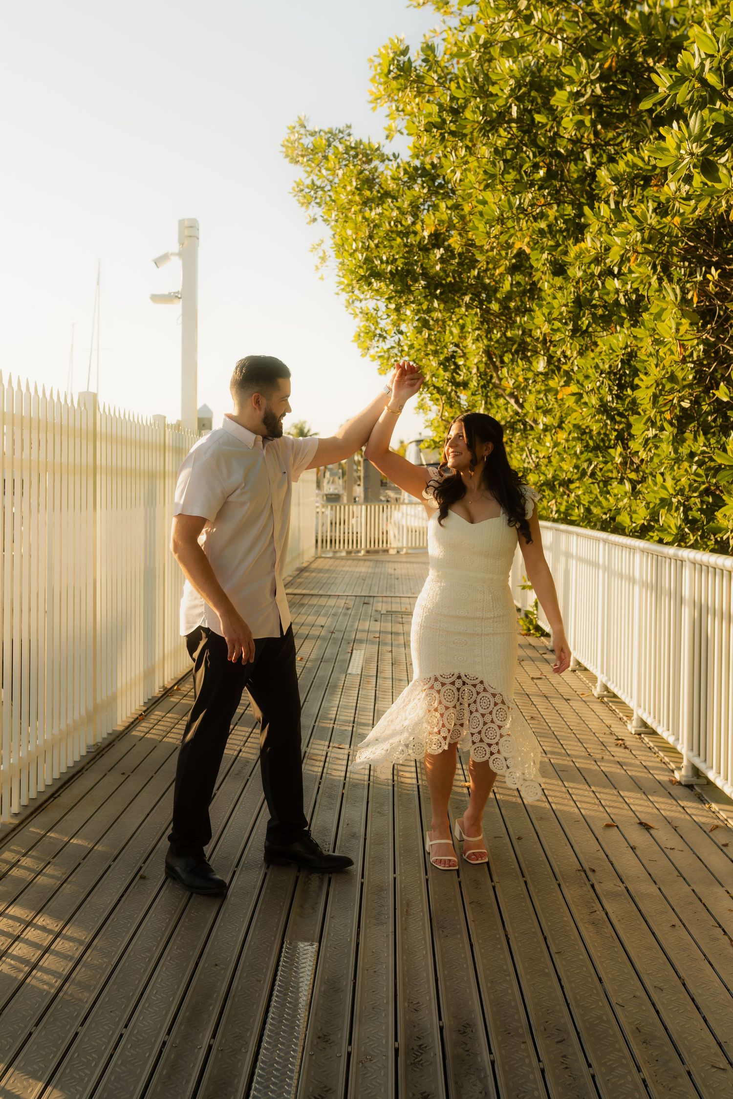 Couple dancing on wooden boardwalk at sunset, man spinning woman in white lace dress.