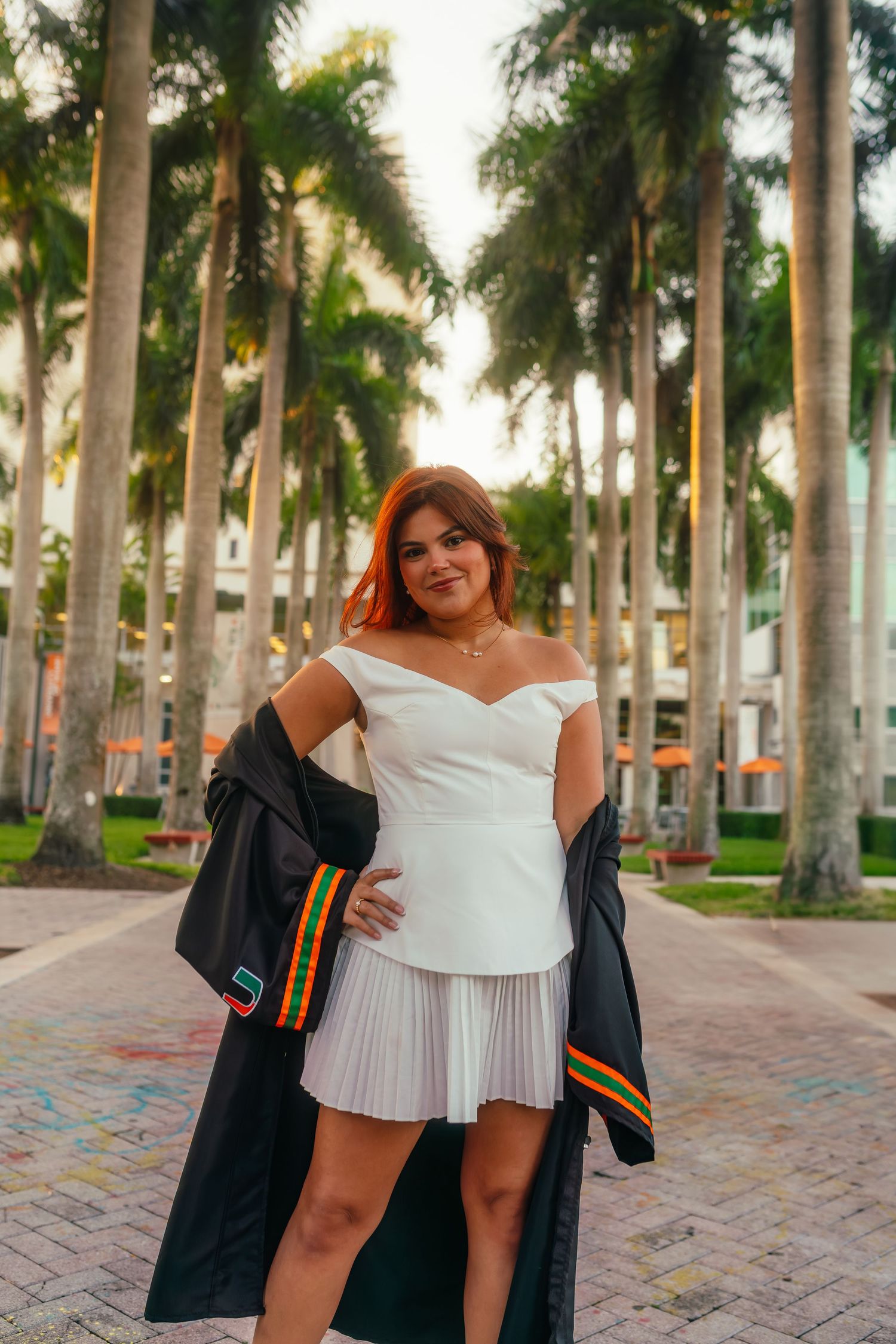 Graduate in white off-shoulder dress holds black gown among tall palm trees.