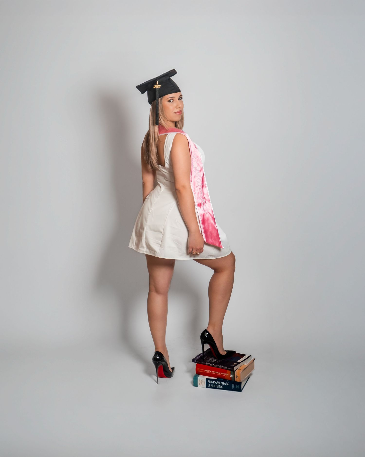 Woman in white dress and graduation cap poses on stacked books wearing black heels.