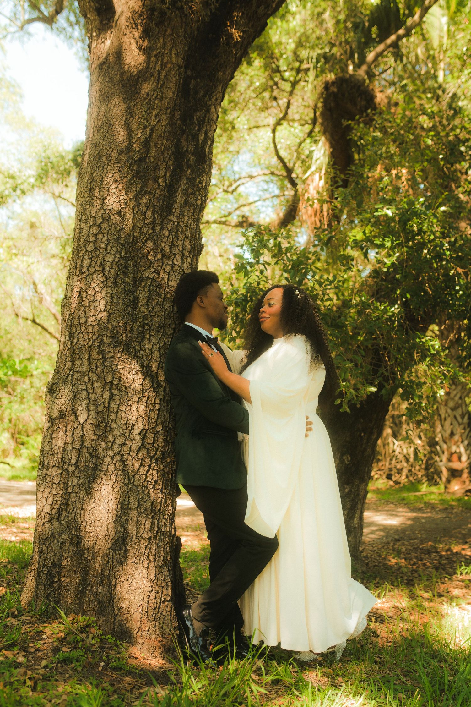 Black couple in wedding attire sharing romantic moment leaning against large tree in sunlit forest.