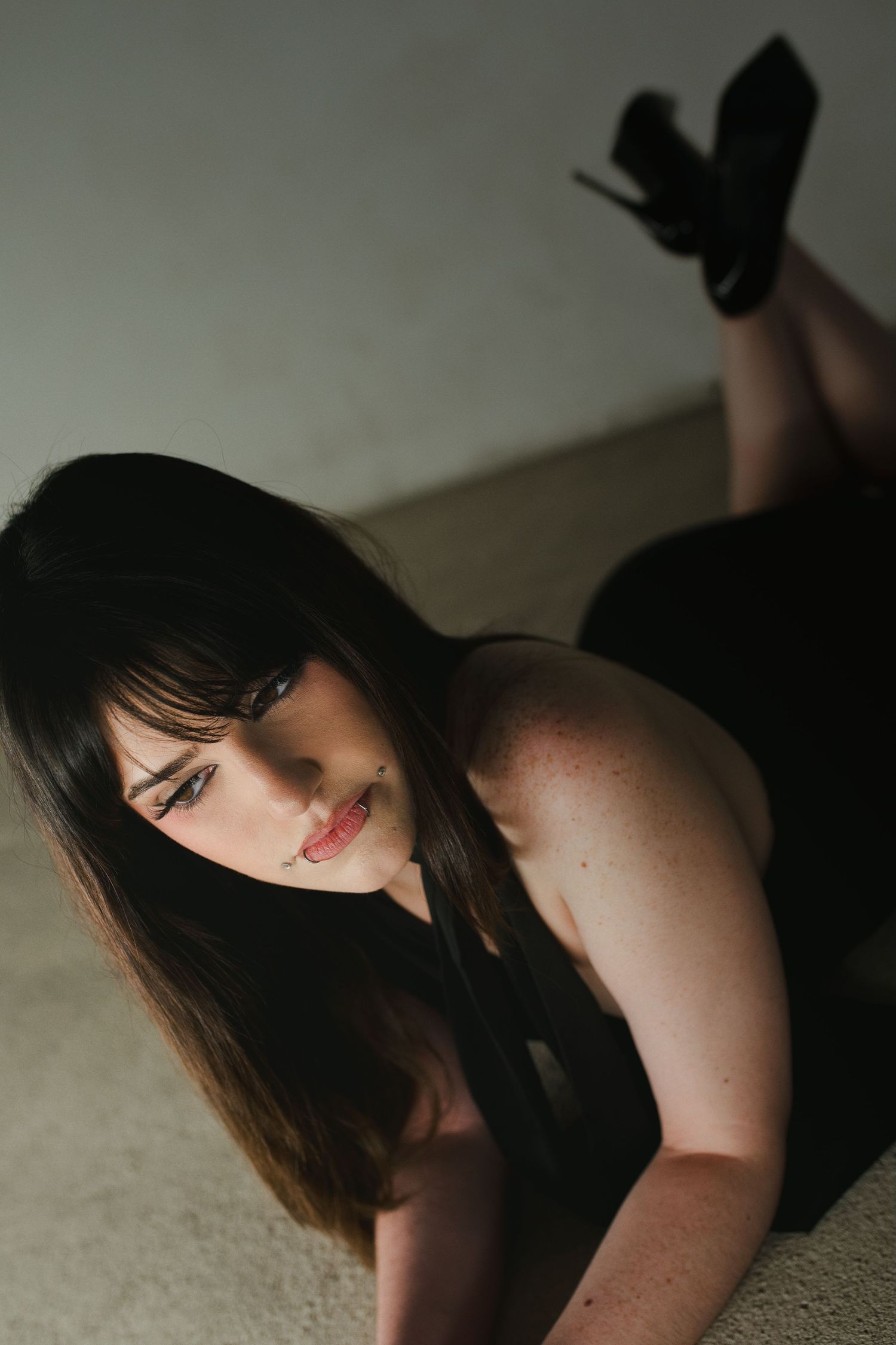 Young woman with dark hair in black outfit and heels posing on floor in dramatic low-light photography.