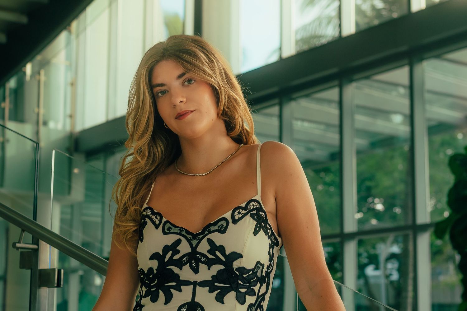 Young woman in elegant black and white floral embroidered dress posing near glass building railing.