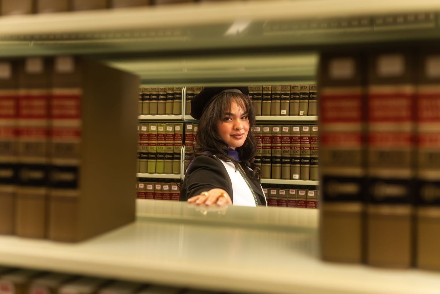 Graduate in cap and gown peers through law library bookshelves filled with legal volumes.