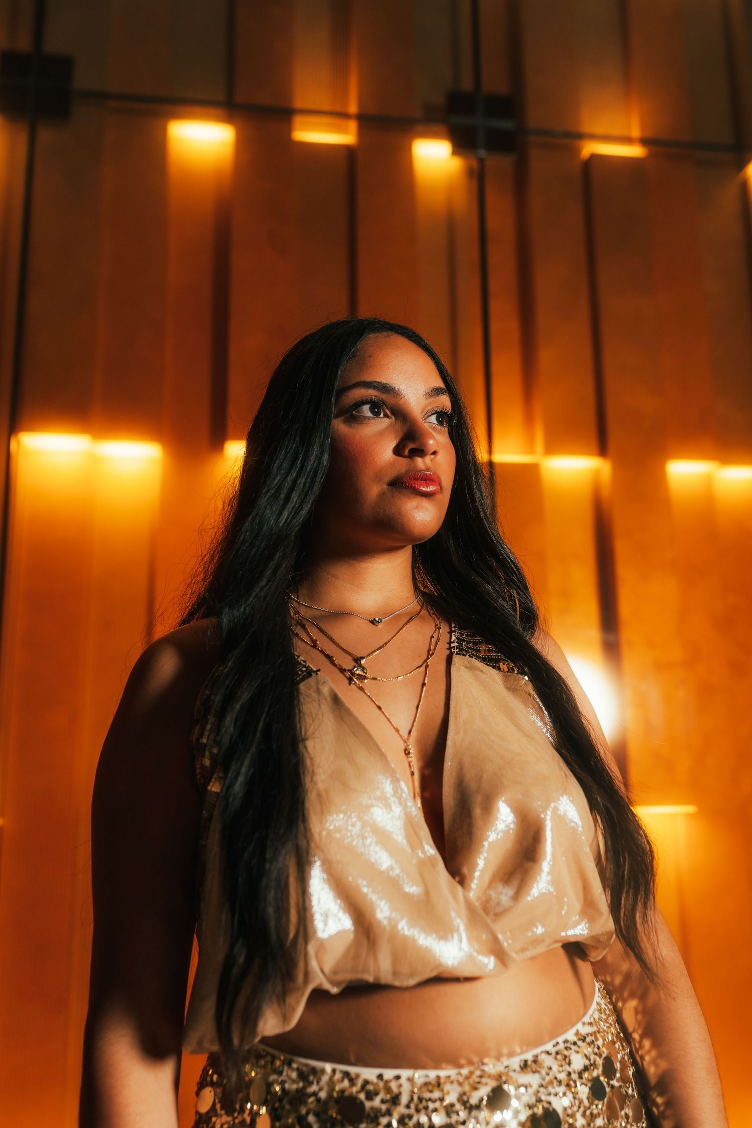 Woman with long dark hair in gold crop top and layered necklaces poses before warm amber backlit wall.