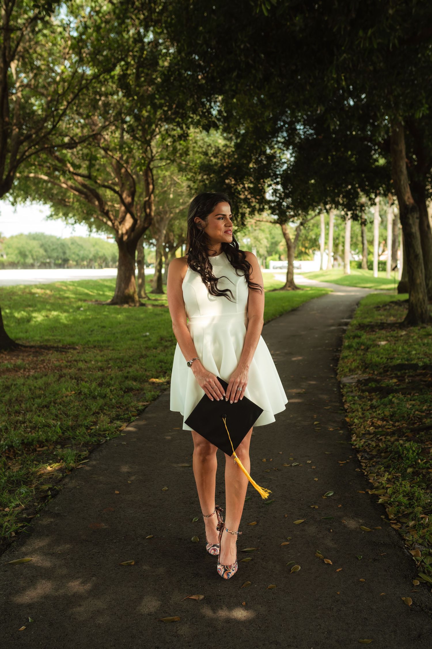 Young woman in white dress holding graduation cap walks along tree-lined park pathway.