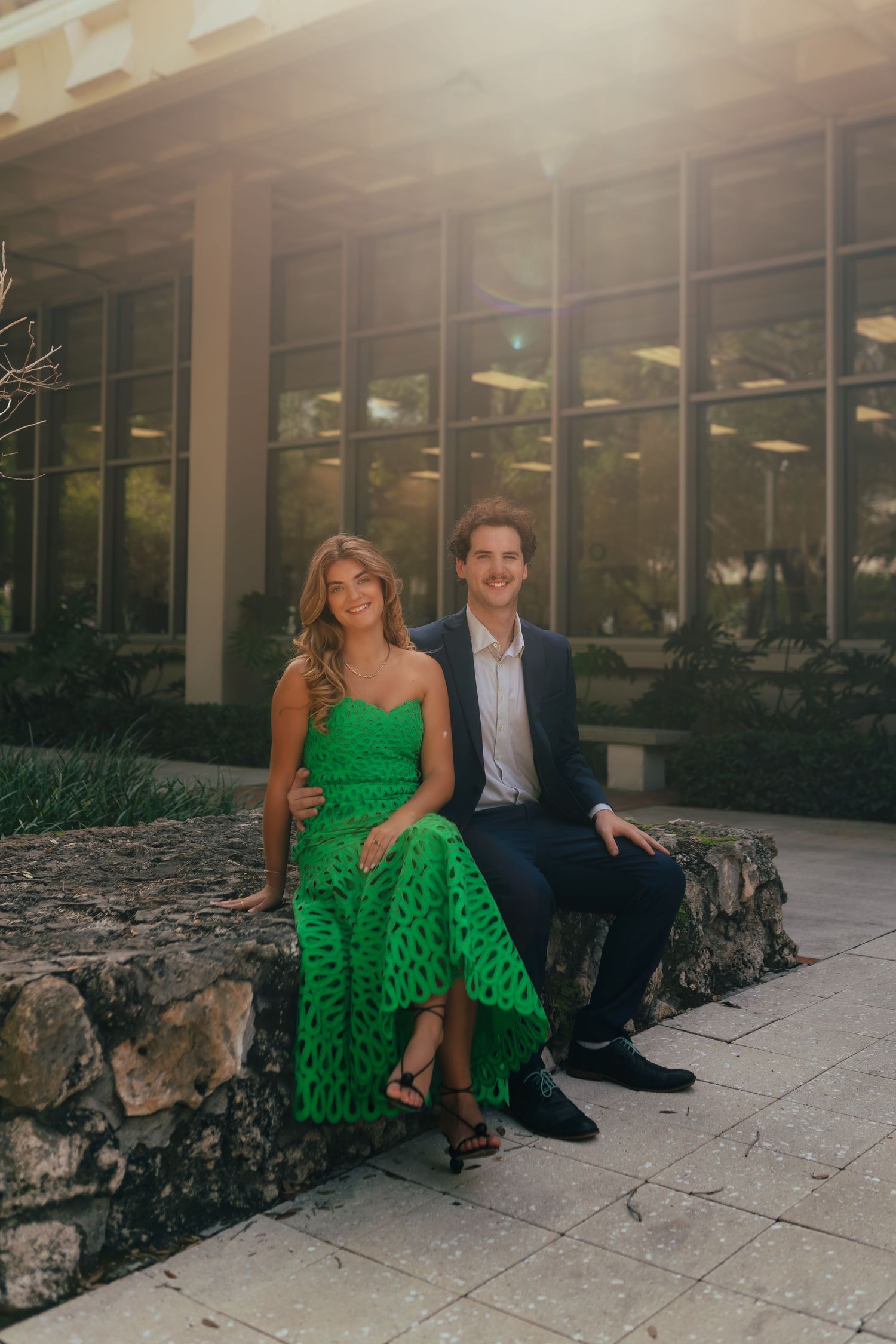 Couple sitting on stone wall outdoors, woman in green dress and man in navy suit, golden hour sunlight behind them.