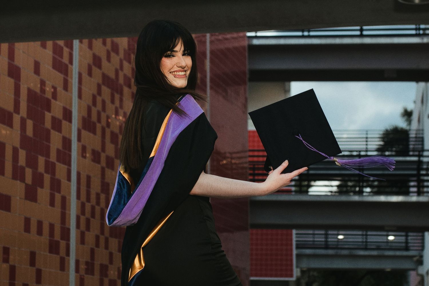 Female graduate in black gown with purple and gold hood holding mortarboard cap outdoors.