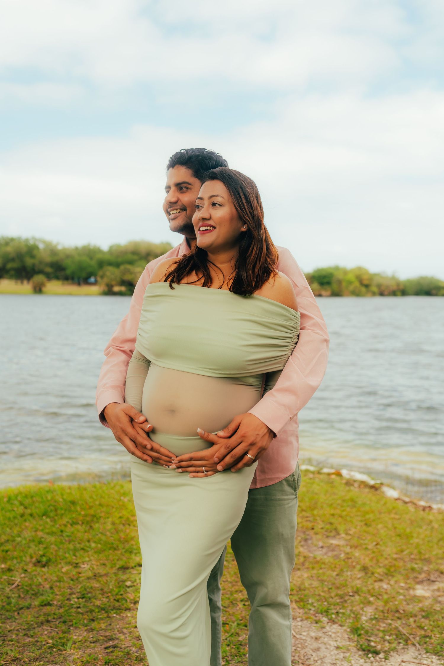 Expecting couple embracing by a lake, man holding pregnant woman's belly from behind, smiling joyfully.