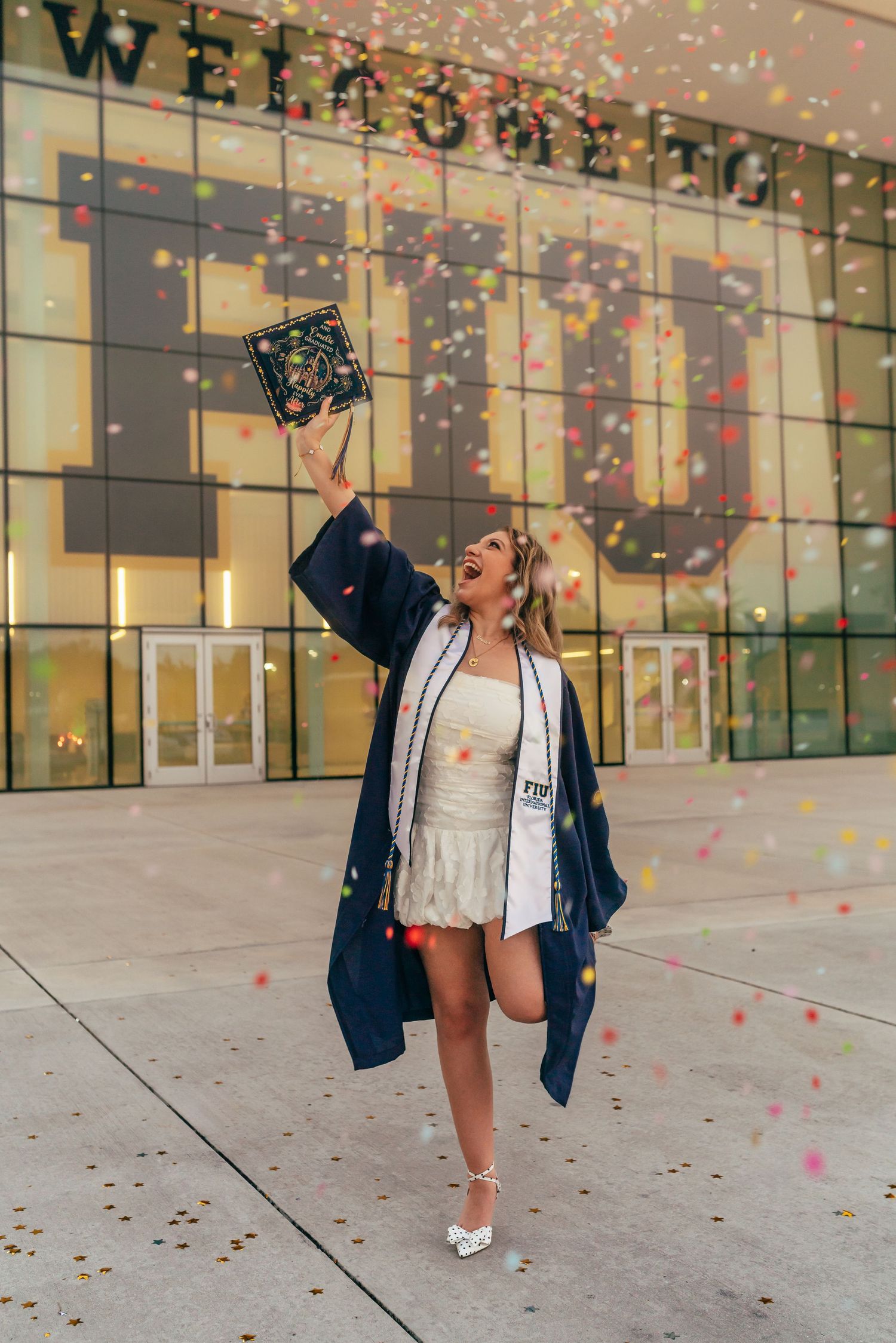 A joyful graduate in a navy gown tosses her cap amid colorful confetti outside an FIU building.