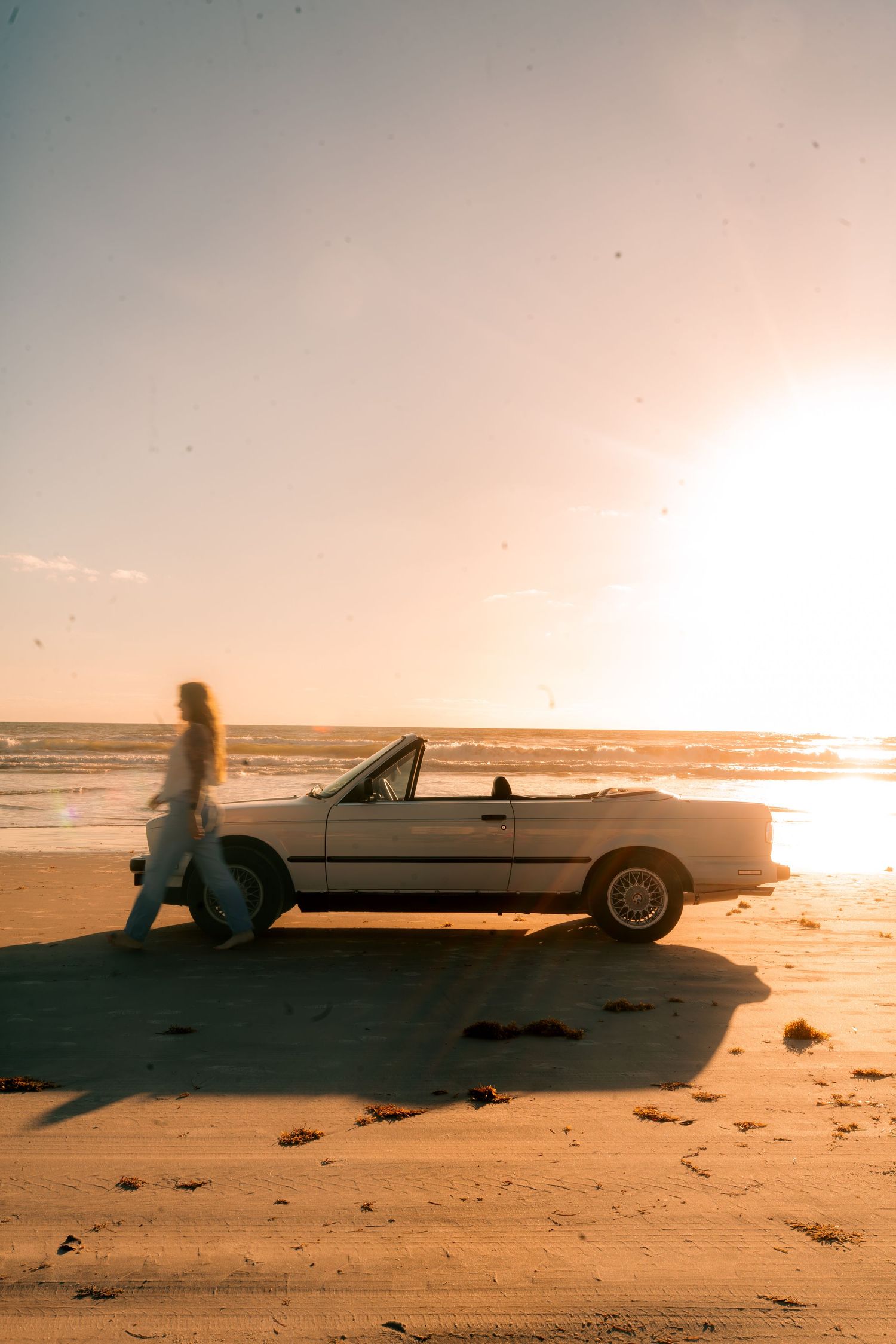 Woman walking beside a white convertible car parked on a sandy beach at golden sunset.