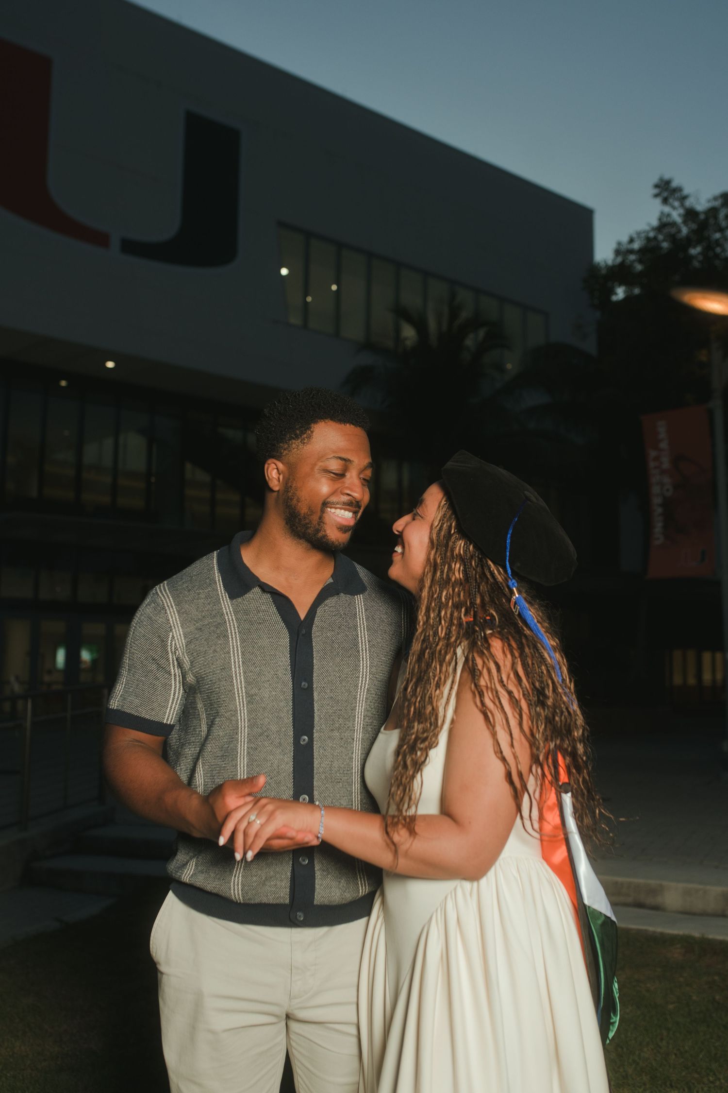 A couple smiles and holds hands at dusk outside the University of Miami, woman wearing graduation cap.