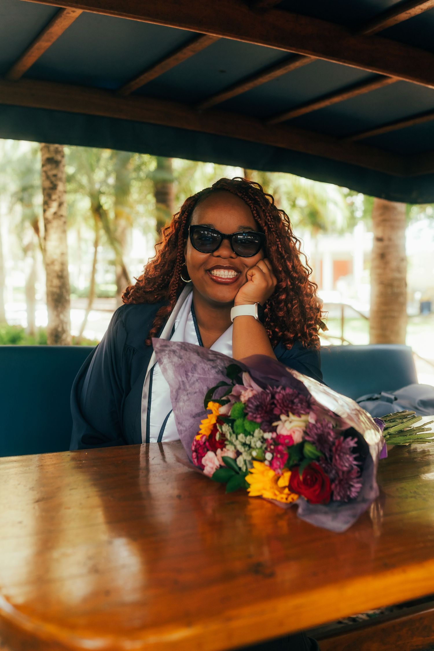 Smiling Black woman with curly hair and sunglasses sitting at a wooden table with a colorful flower bouquet.