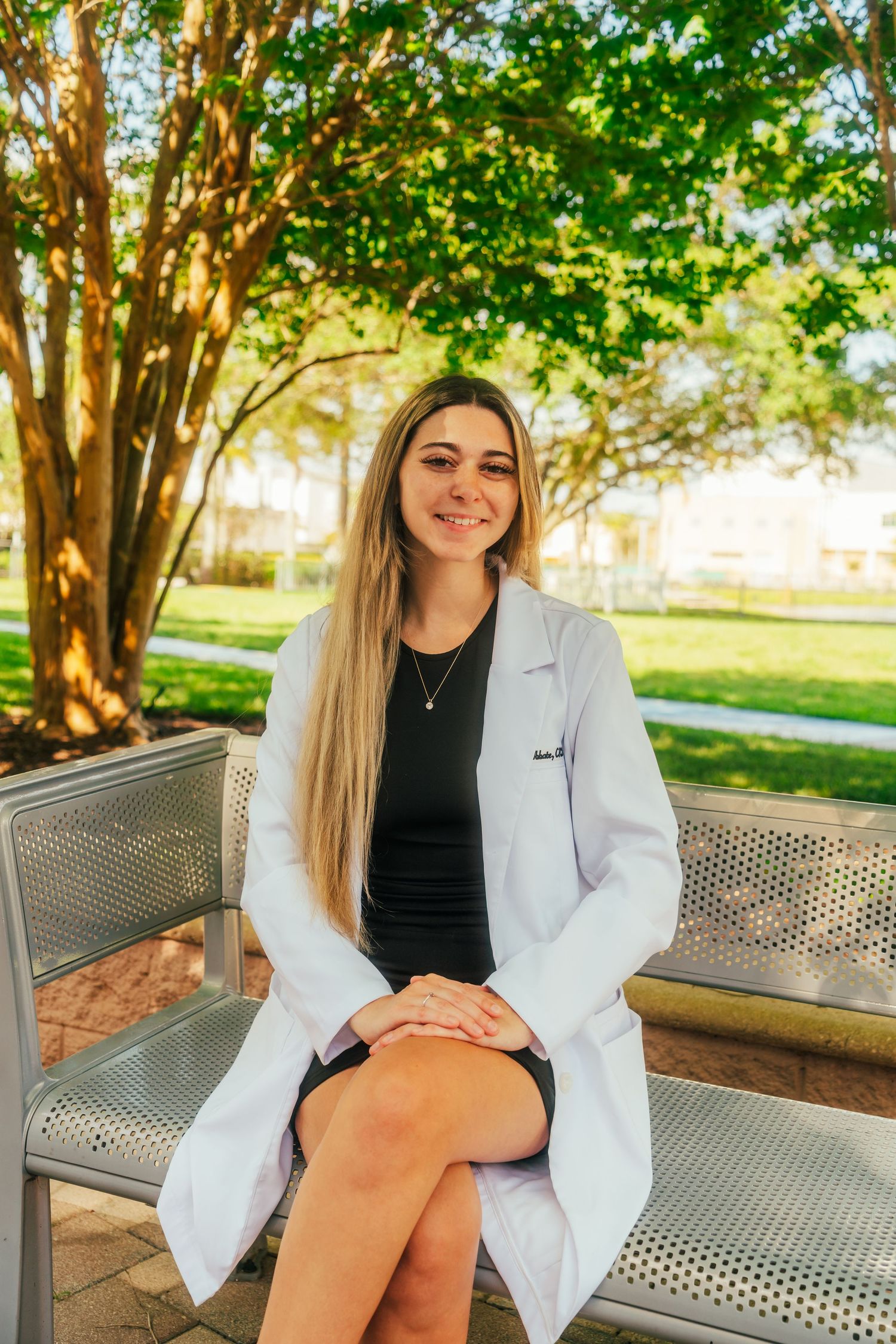 Smiling female medical professional in white lab coat sitting on bench outdoors near green trees on sunny day.