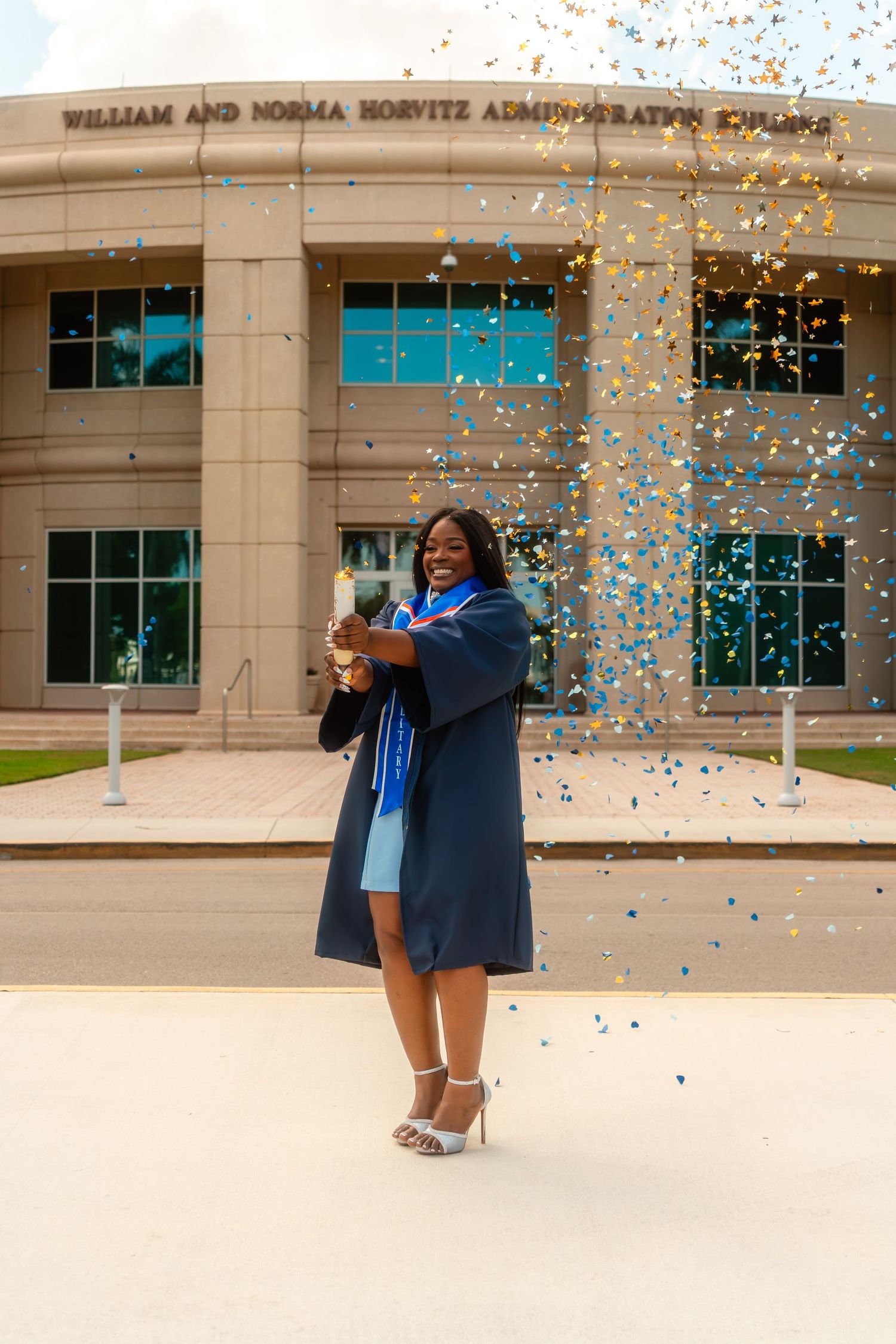 Graduate in navy gown celebrates with confetti outside William and Norma Horvitz building.