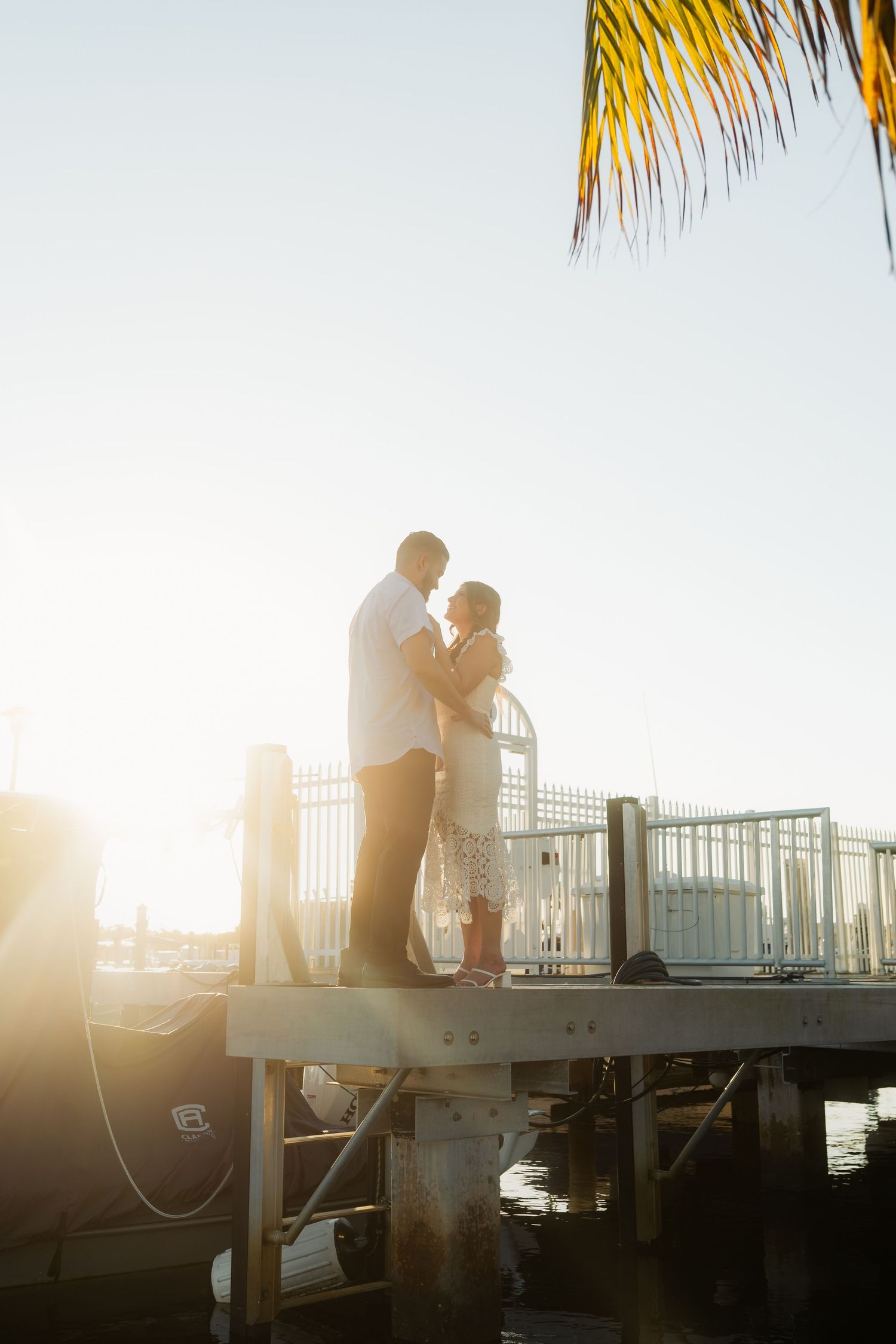 Couple silhouetted against bright sunset light standing together on waterfront dock near marina.
