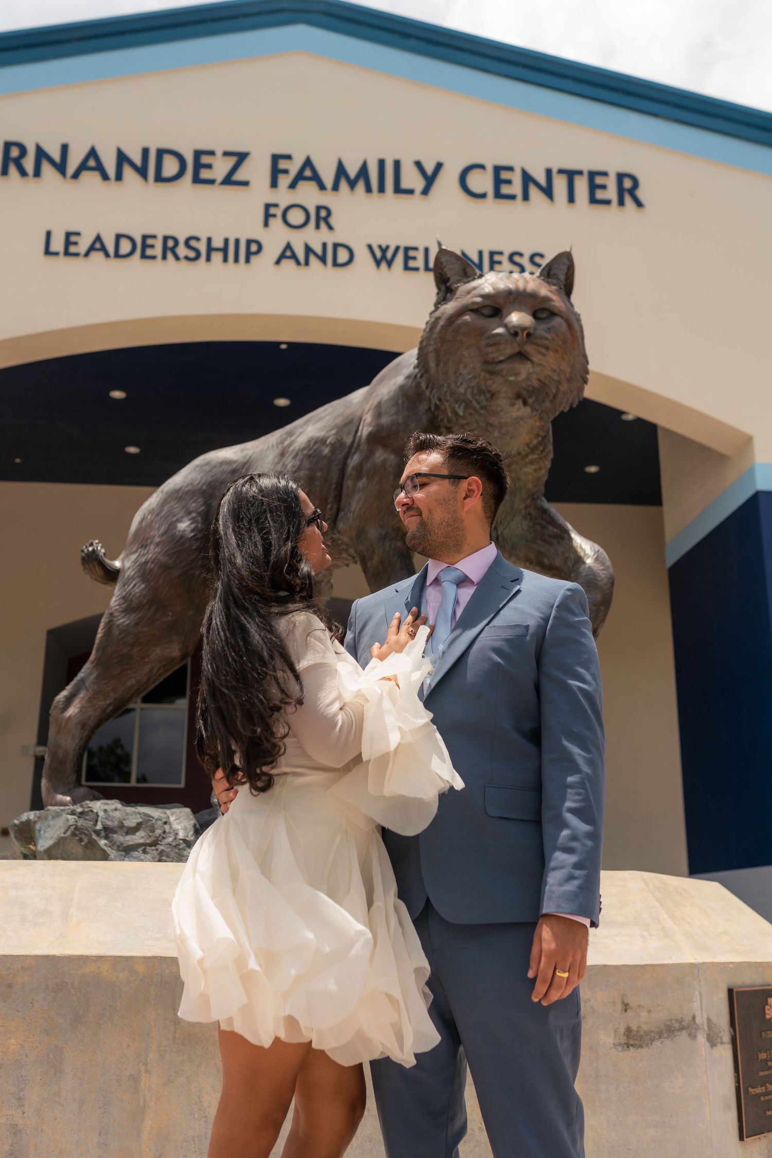 Couple poses romantically in front of Hernandez Family Center for Leadership and Wellness with a wildcat statue.