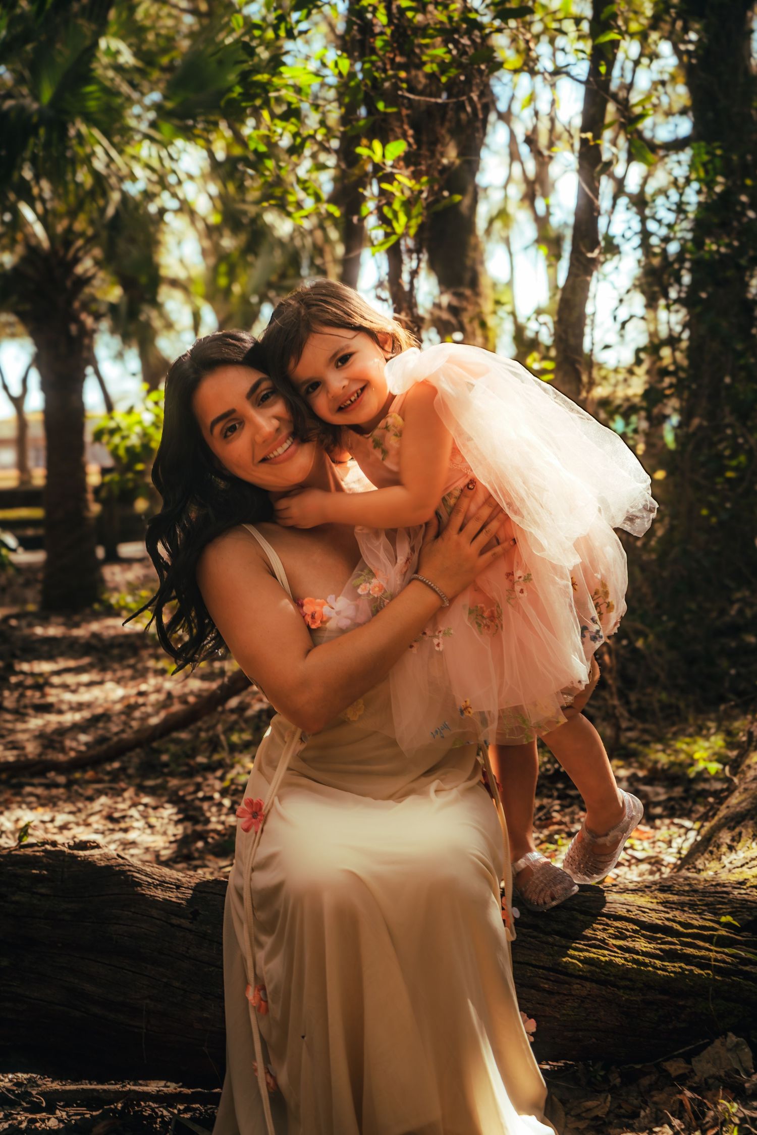 A mother in a floral dress holds her young daughter in a pink tutu outdoors in a sunlit forest setting.