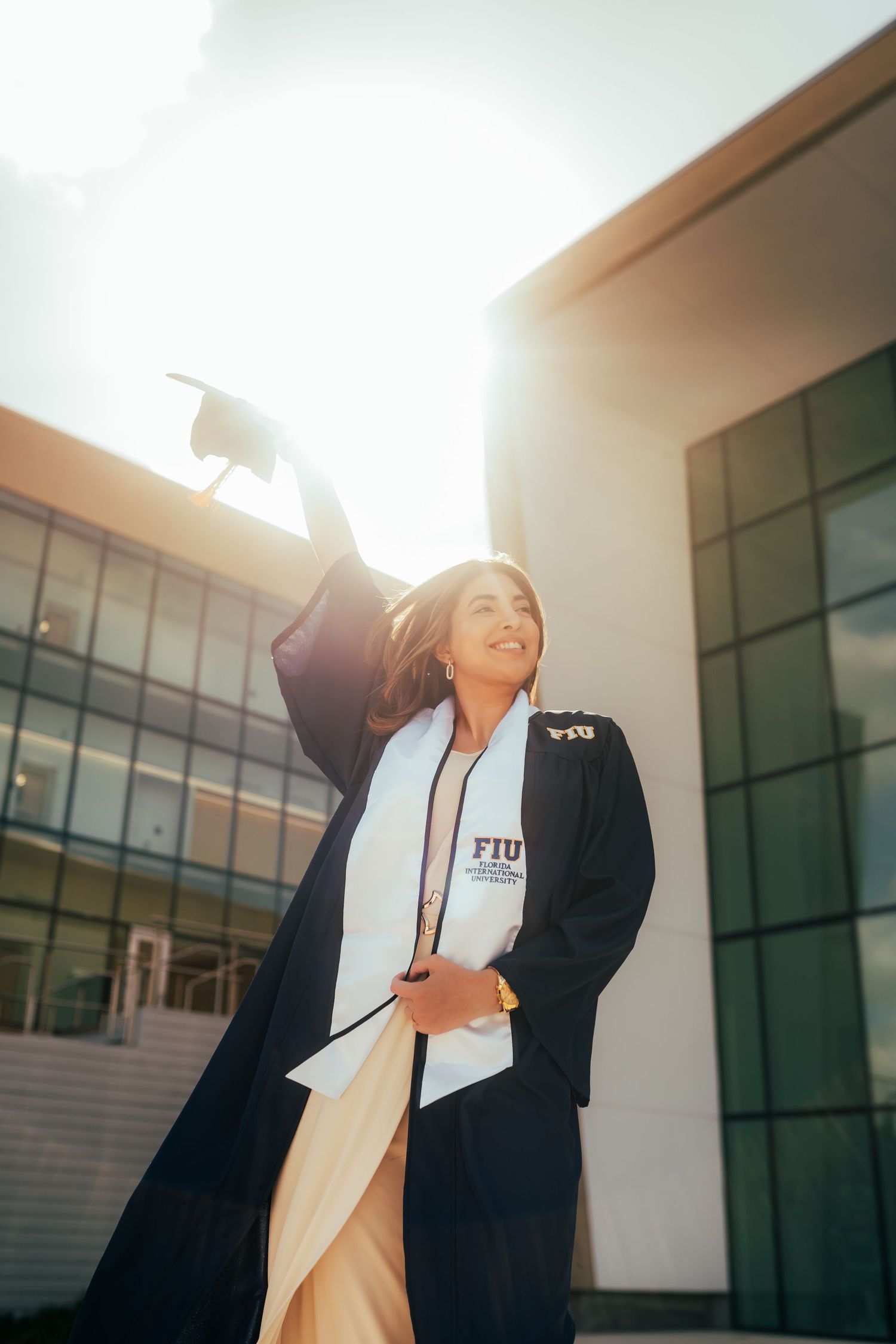 FIU graduate in black cap and gown raises mortarboard triumphantly outside university building in golden sunlight.