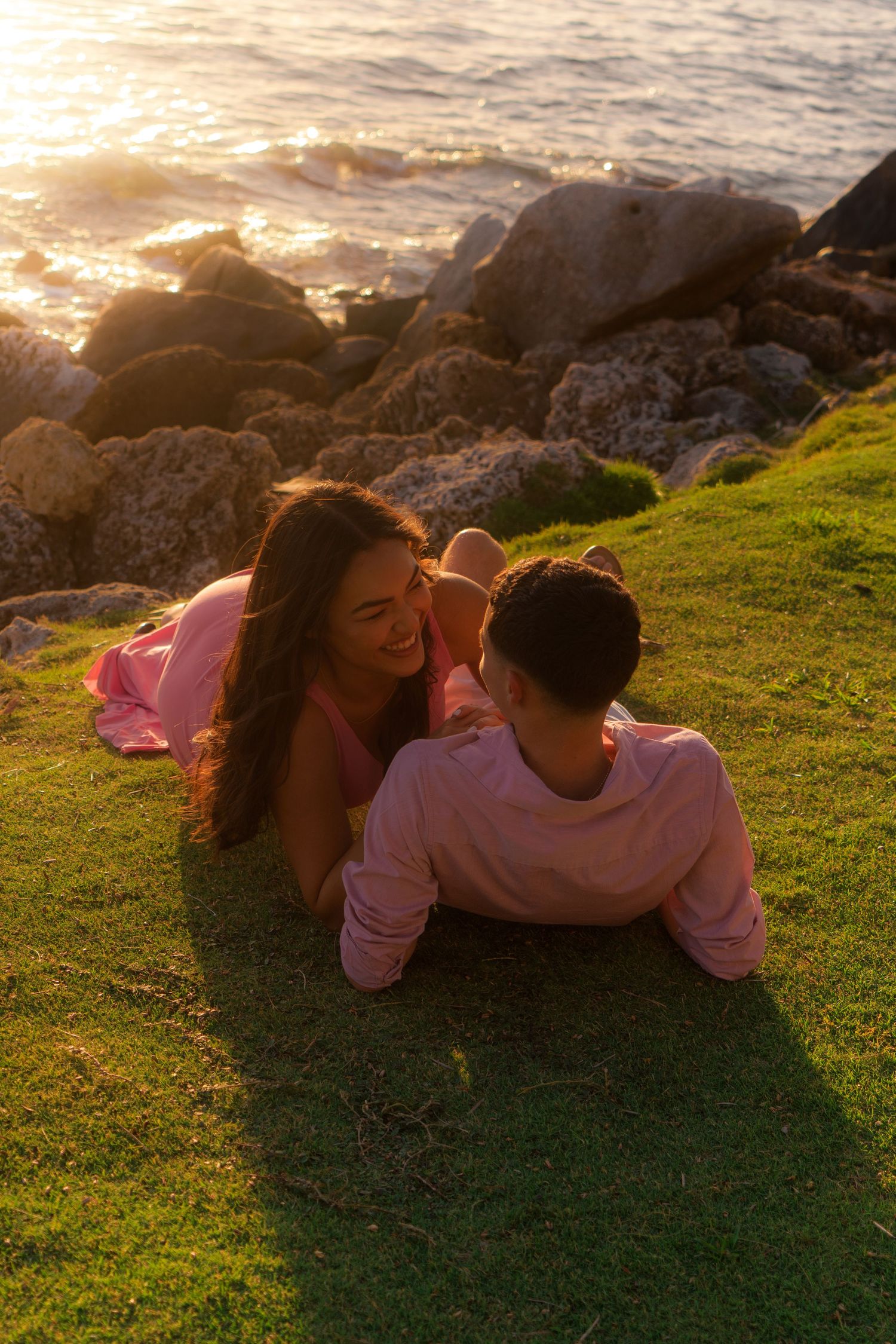Couple lying on green grass by rocky ocean shore during warm golden sunset light.