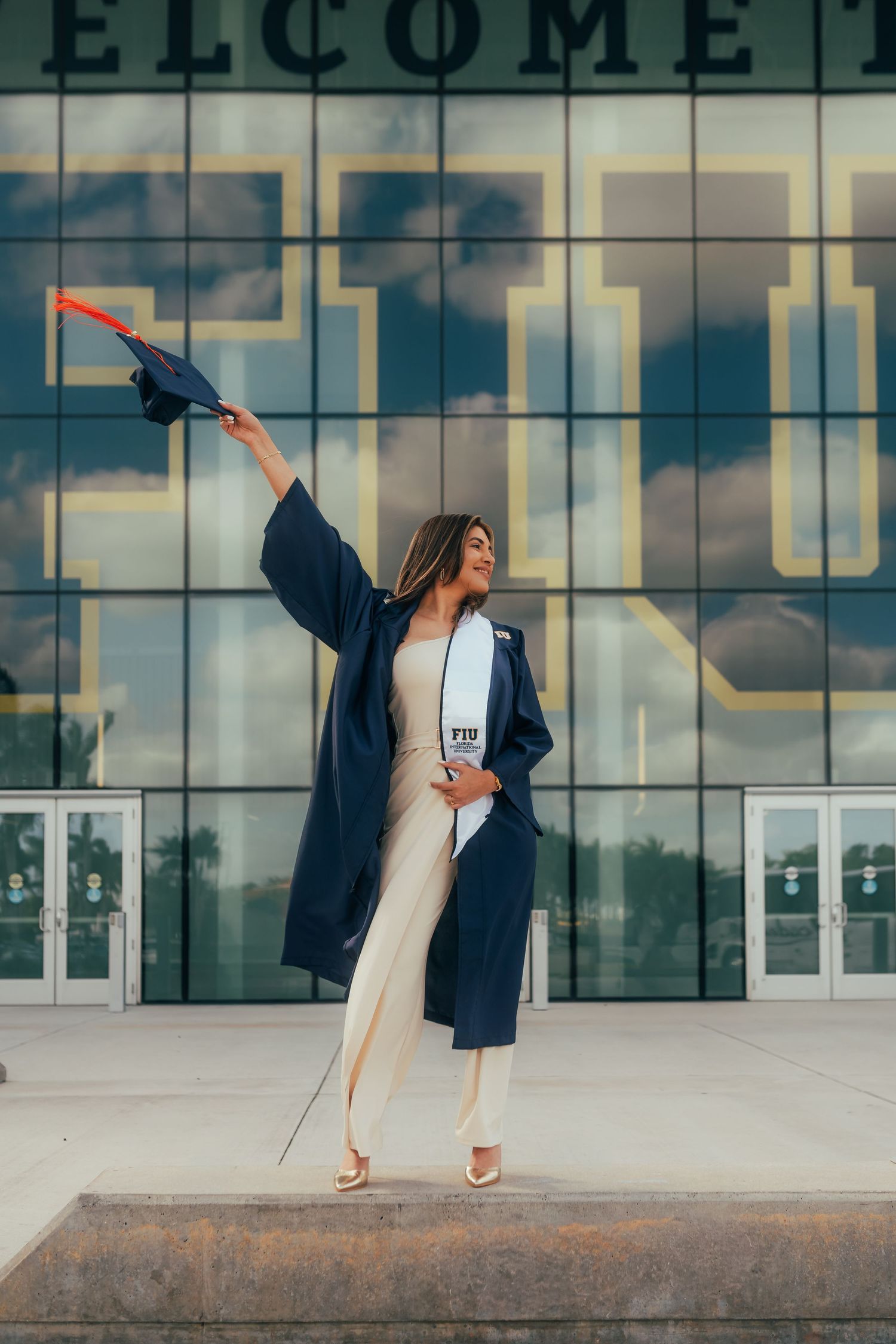 A joyful female graduate in navy cap and gown holds her cap aloft outside FIU university building.