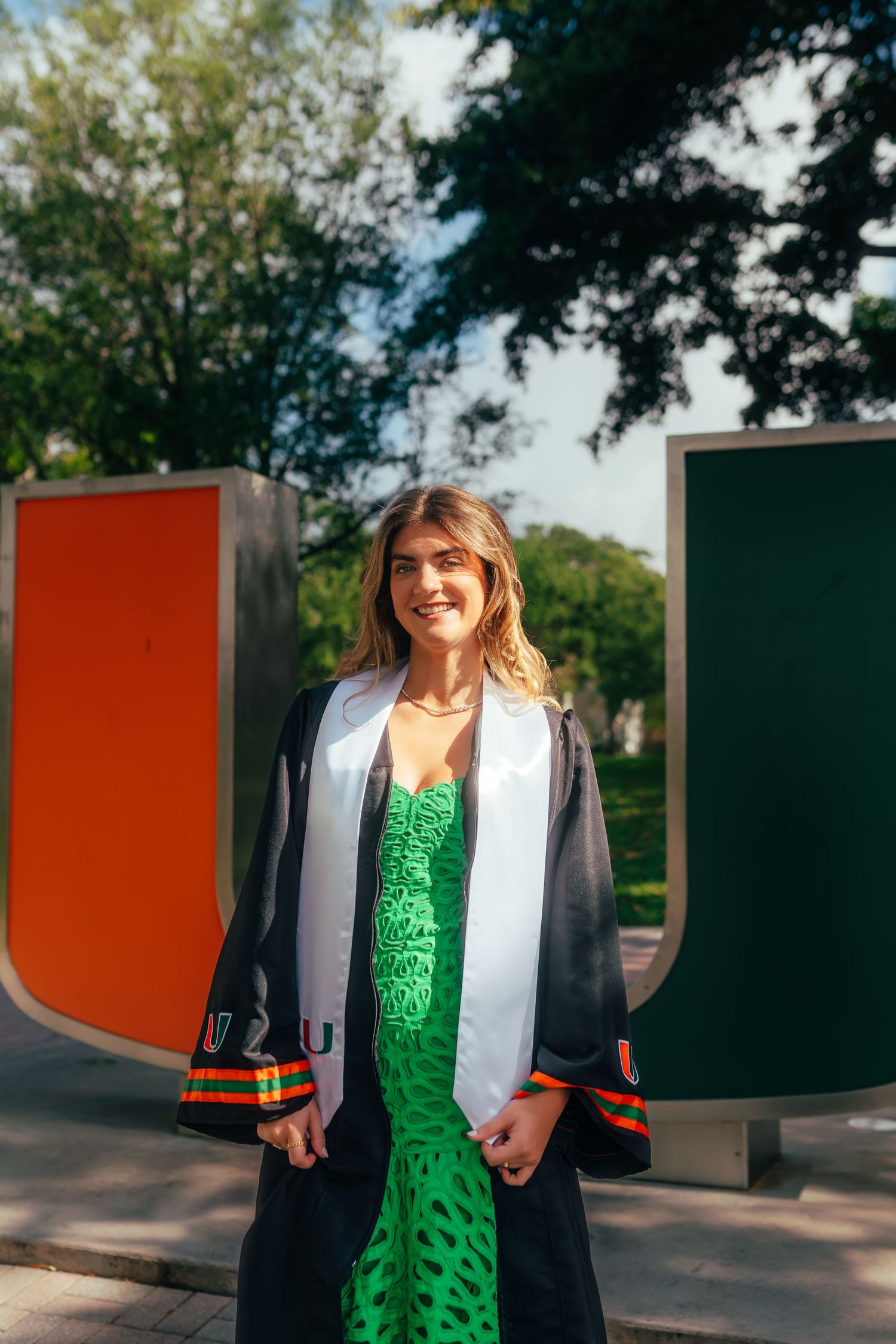 Smiling graduate in black gown and green dress poses in front of University of Miami U sculpture.