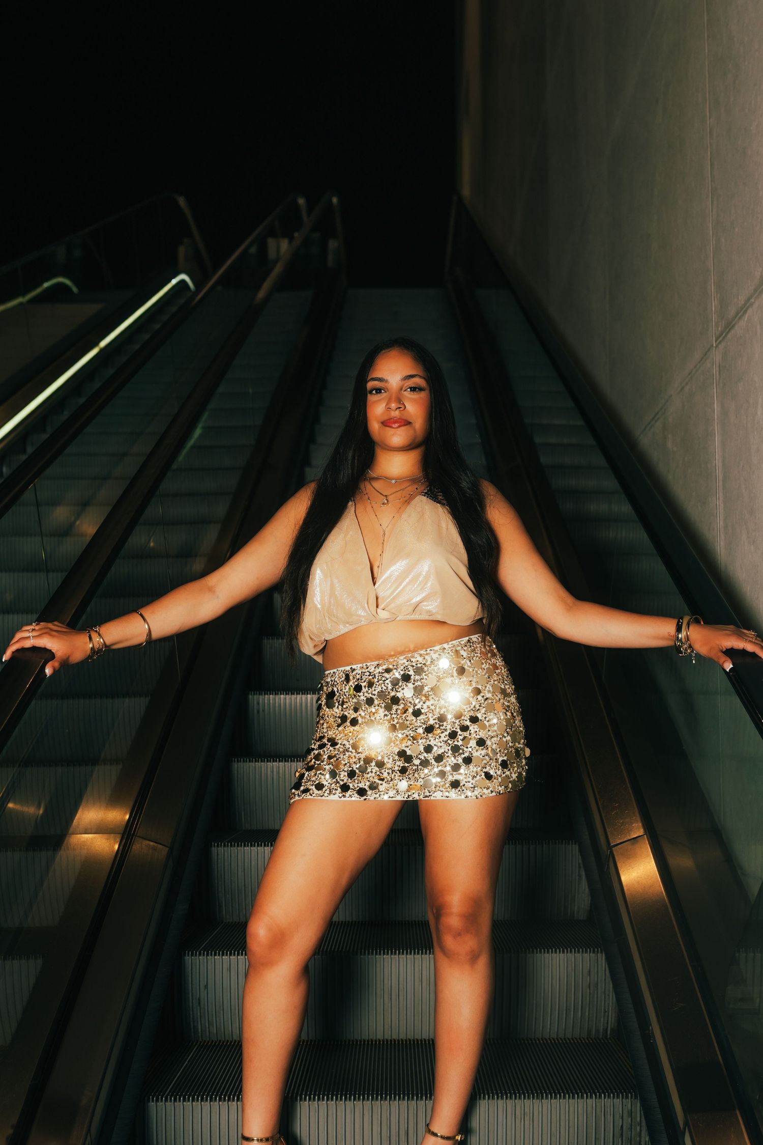Woman in sequin mini skirt and crop top posing confidently on escalator at night.