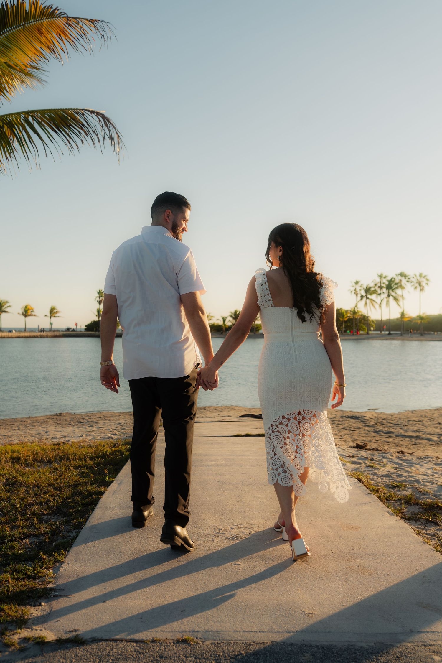 Couple holding hands walking toward tropical waterfront at golden sunset, woman in white lace dress.
