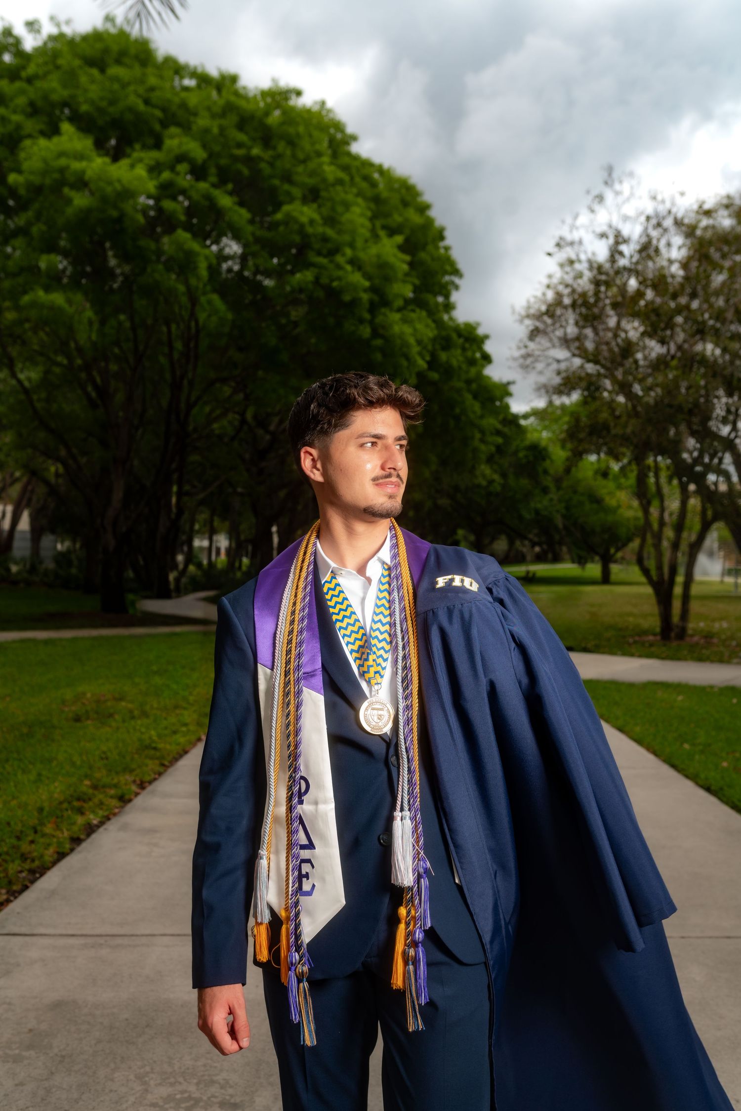 Male graduate in navy gown with purple stole and medals poses on campus pathway.