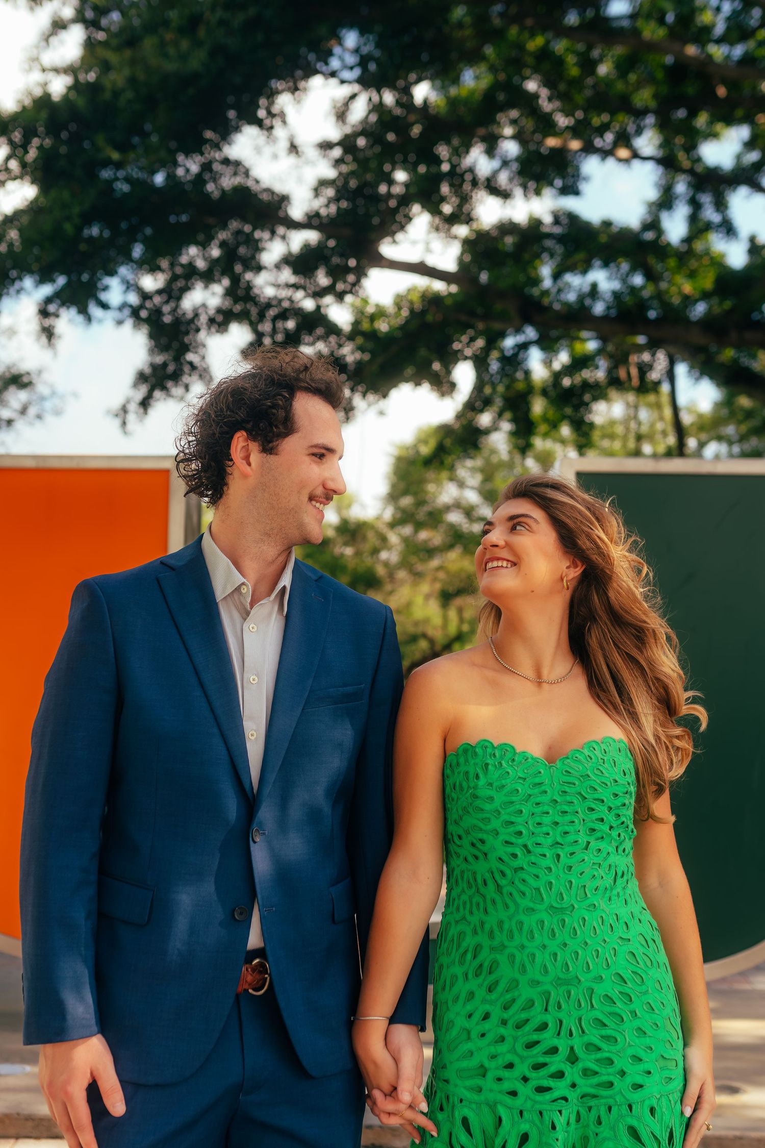 A smiling couple holds hands outdoors, man in blue suit and woman in green strapless dress, with colorful walls behind.