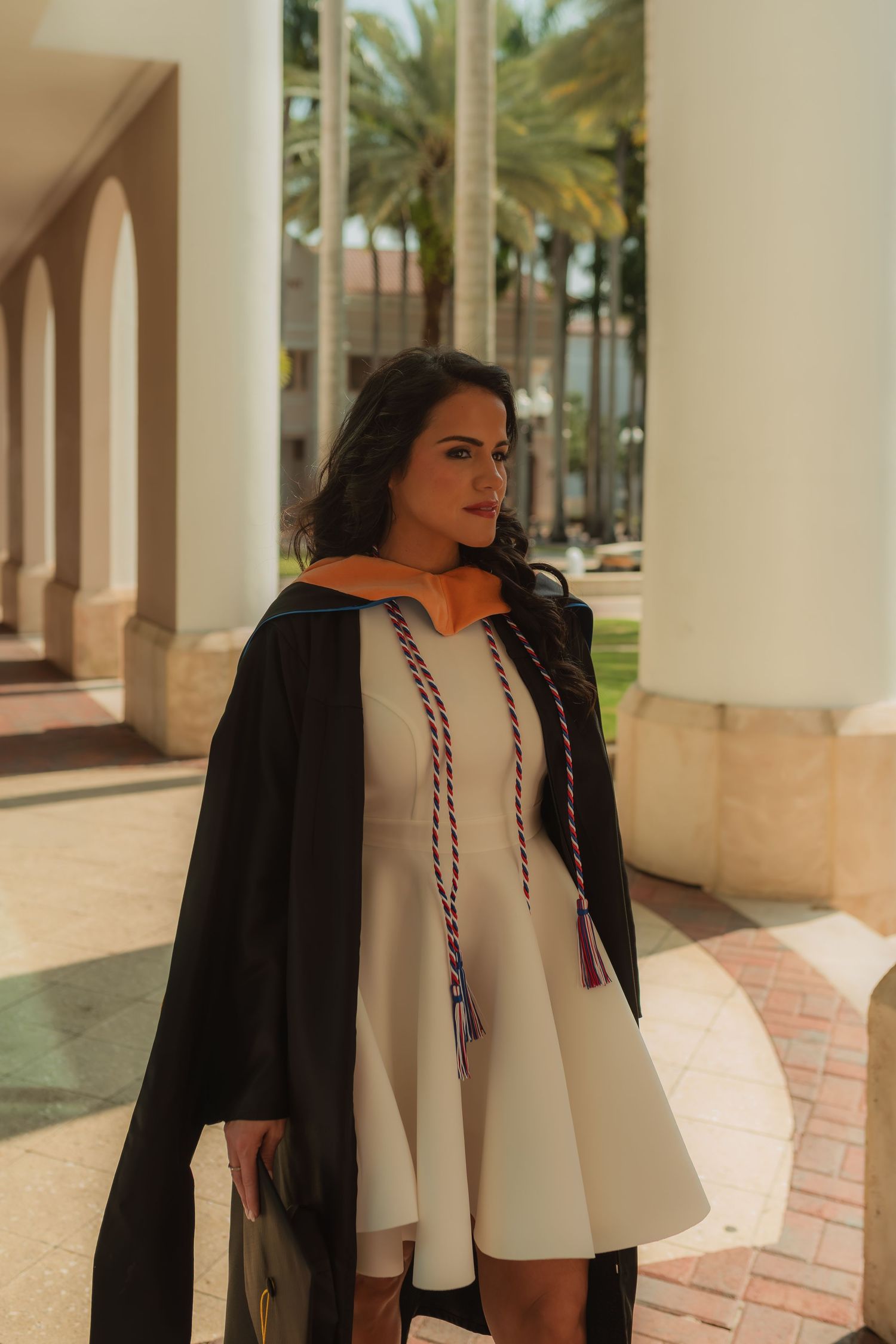 Female graduate in black gown with honor cords standing between white columns on campus.