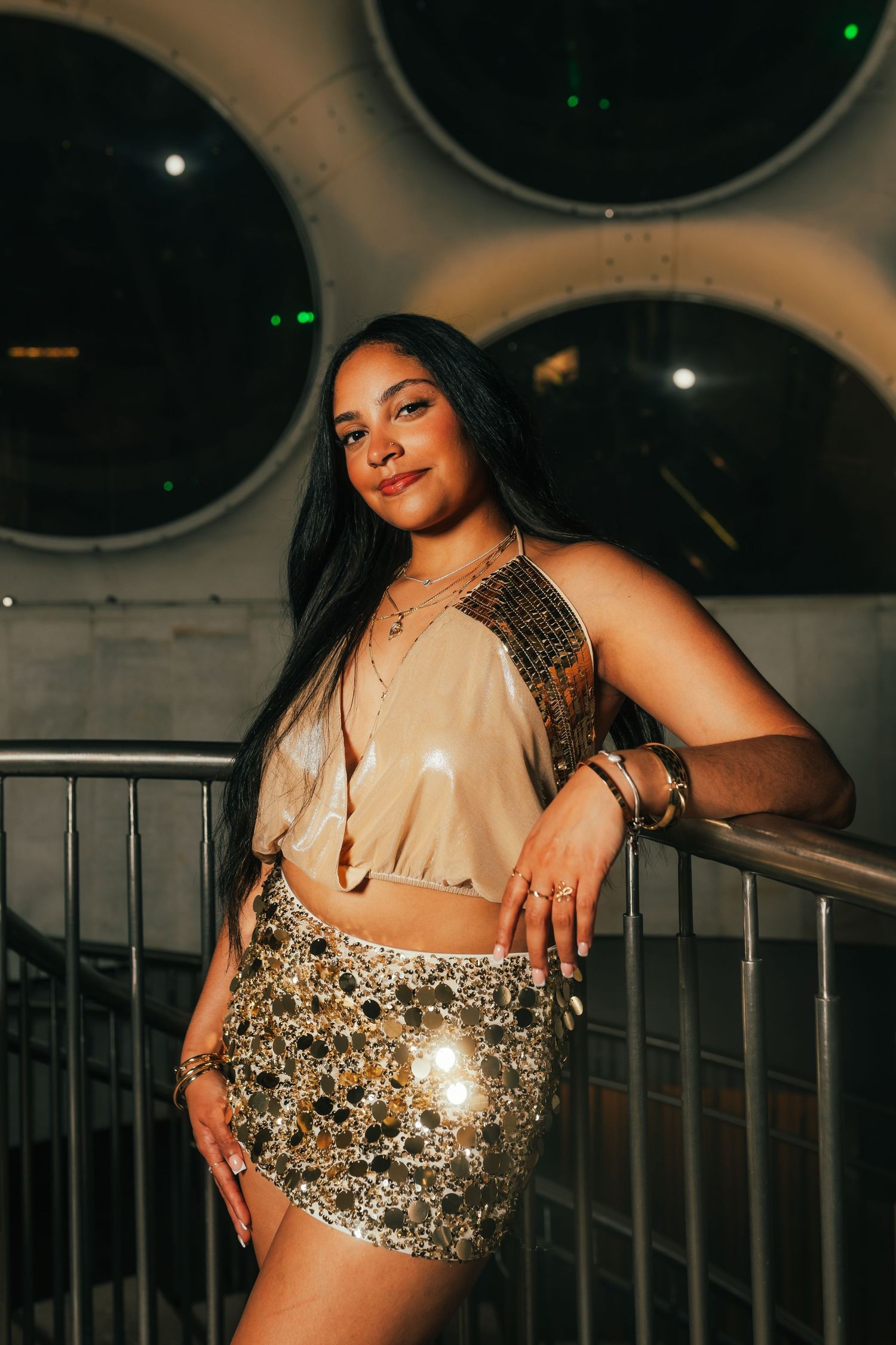 A young woman in a gold sequin skirt and beige crop top poses confidently by a railing in a dimly lit venue.