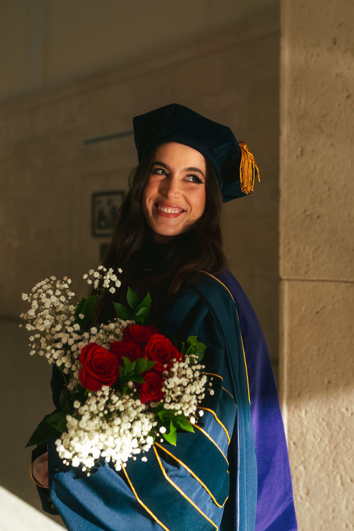 Smiling graduate in teal doctoral gown holds bouquet of red roses and white flowers.