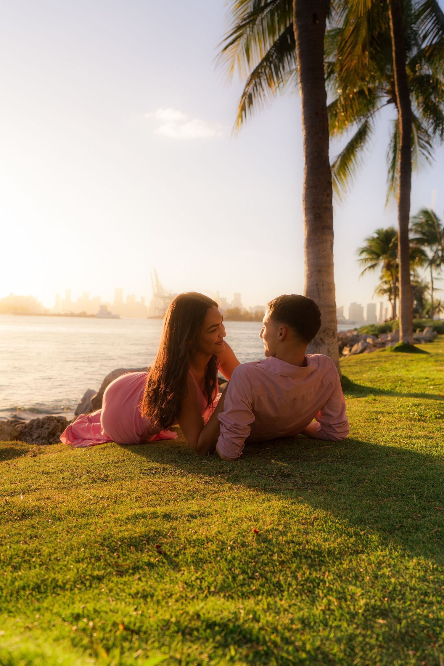 Couple in pink outfits lying on grass by the waterfront at sunset with palm trees and city skyline.