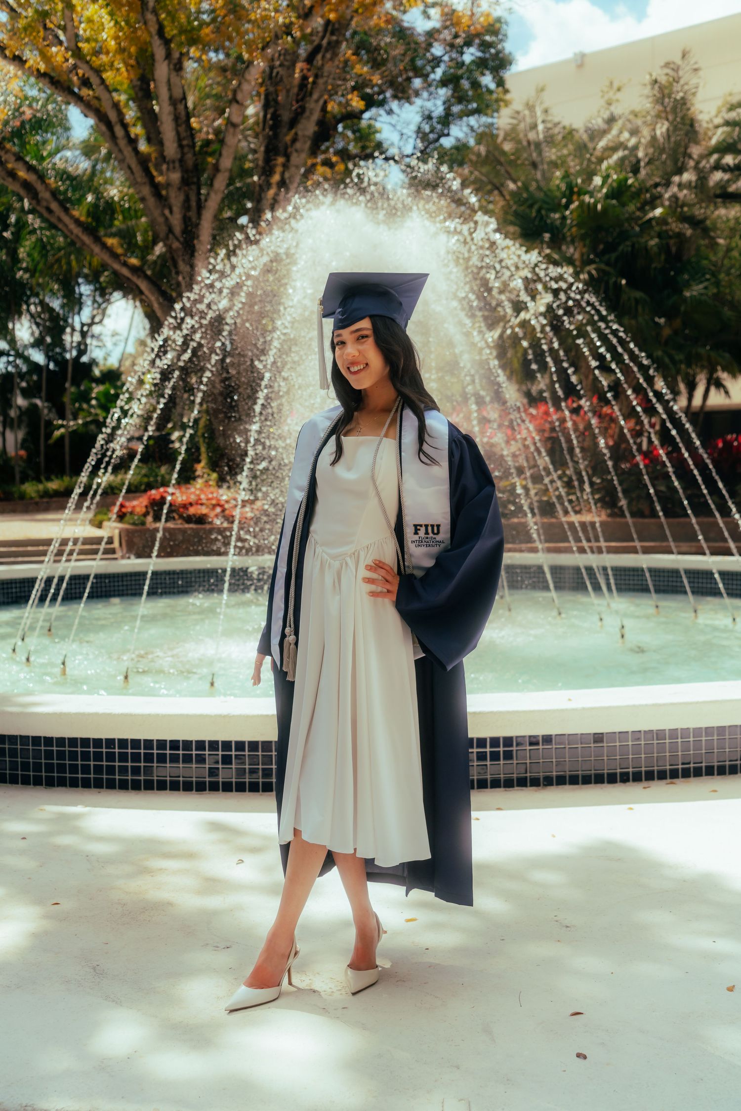 FIU graduate in cap and gown poses in front of a fountain on campus, wearing a white dress and heels.