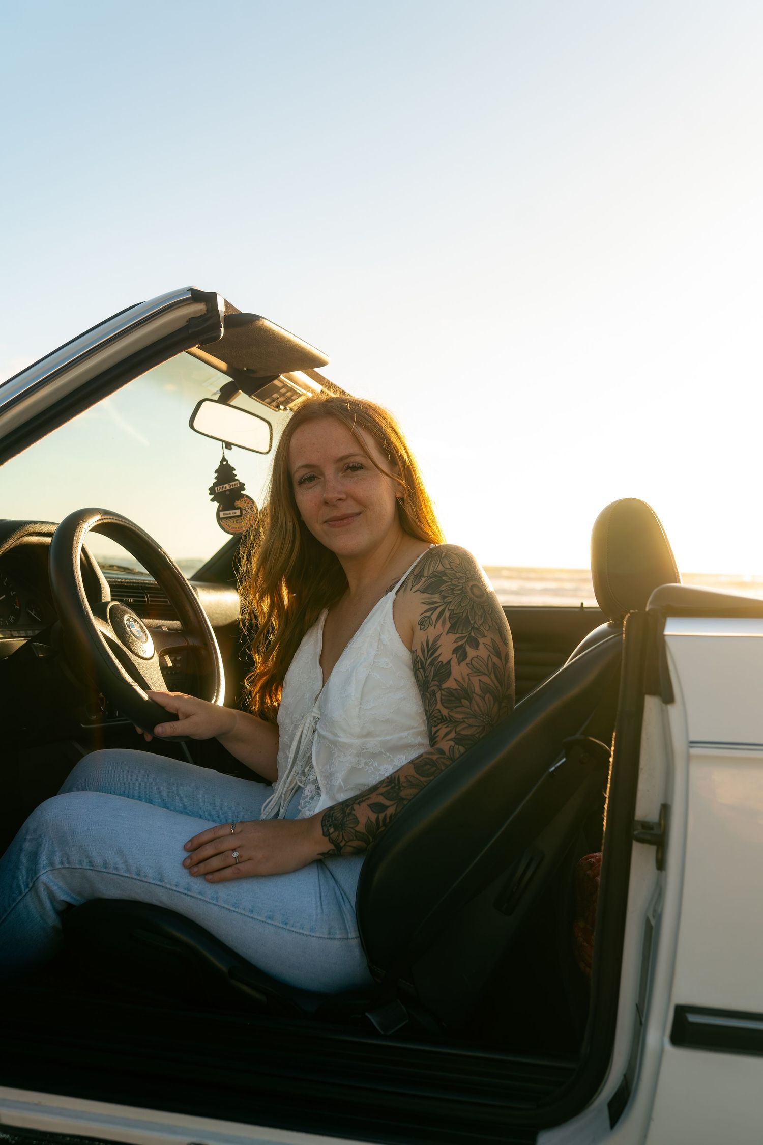 Young woman with sleeve tattoo sits in white convertible car at sunset, wearing white top and jeans.