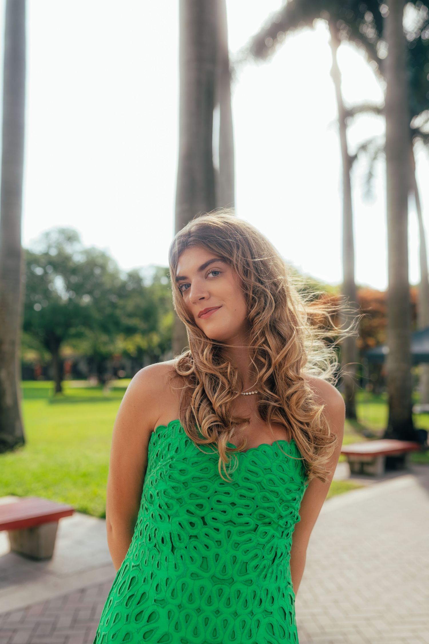 Young woman in green strapless dress posing outdoors in tropical park with palm trees on sunny day.