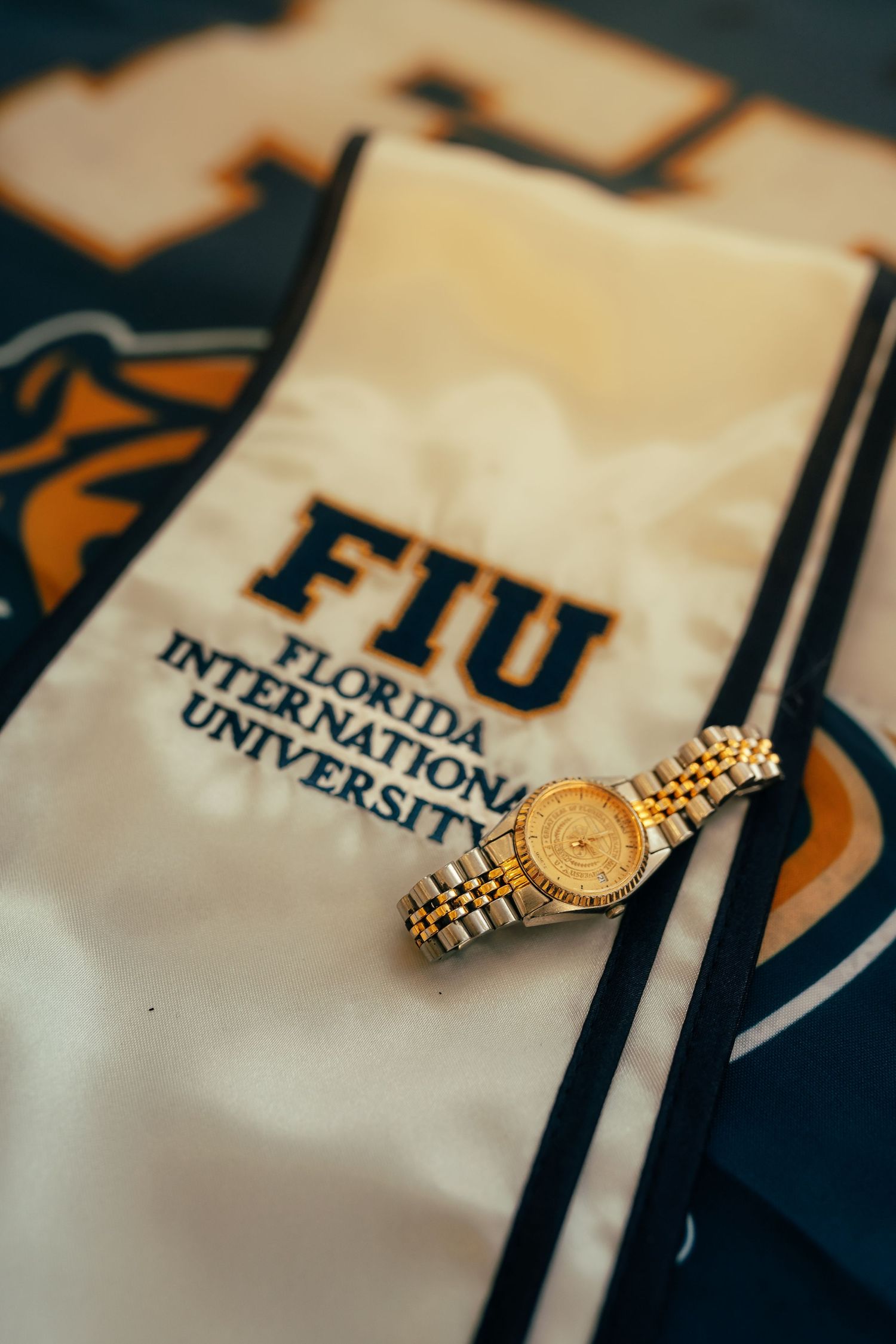 Gold wristwatch resting on an FIU Florida International University white satin sash with university apparel in background.