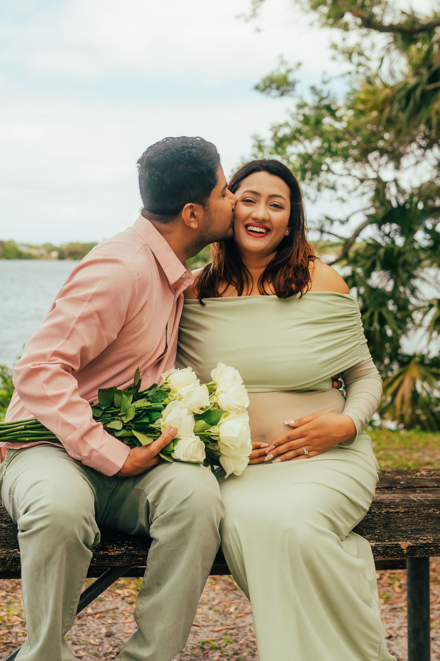 Pregnant woman in sage dress holding white roses receives a cheek kiss from partner on a bench.